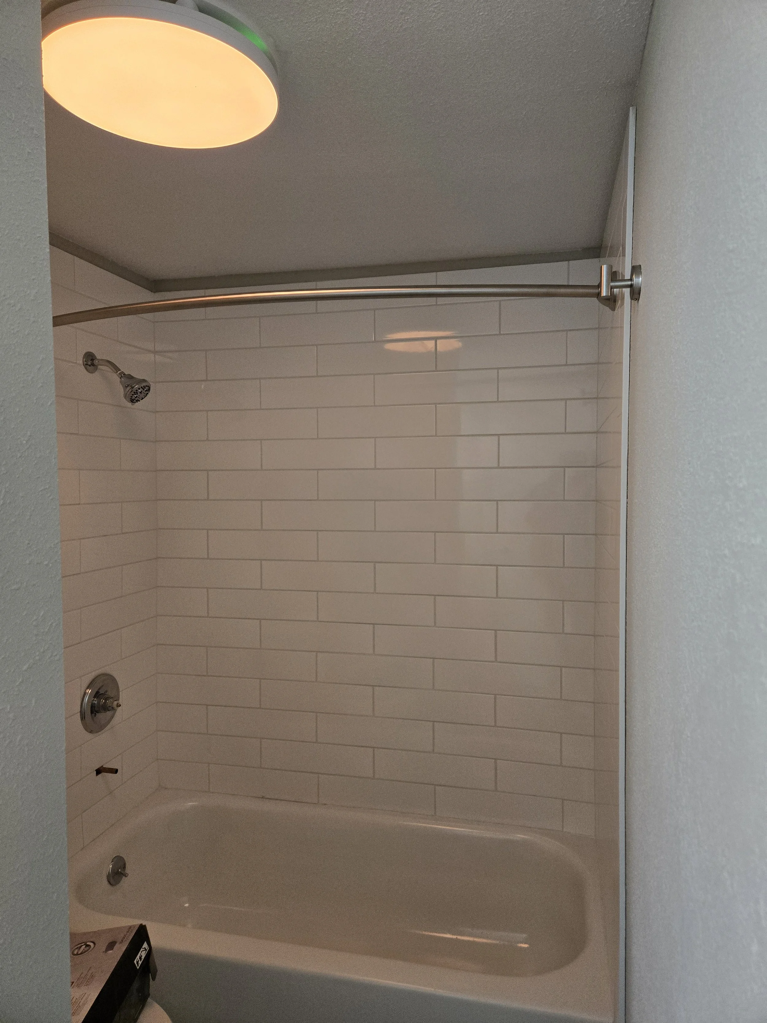 Bathroom shower with white subway tiles, a curved shower curtain rod, a showerhead, and a bathtub. There is a ceiling light fixture.