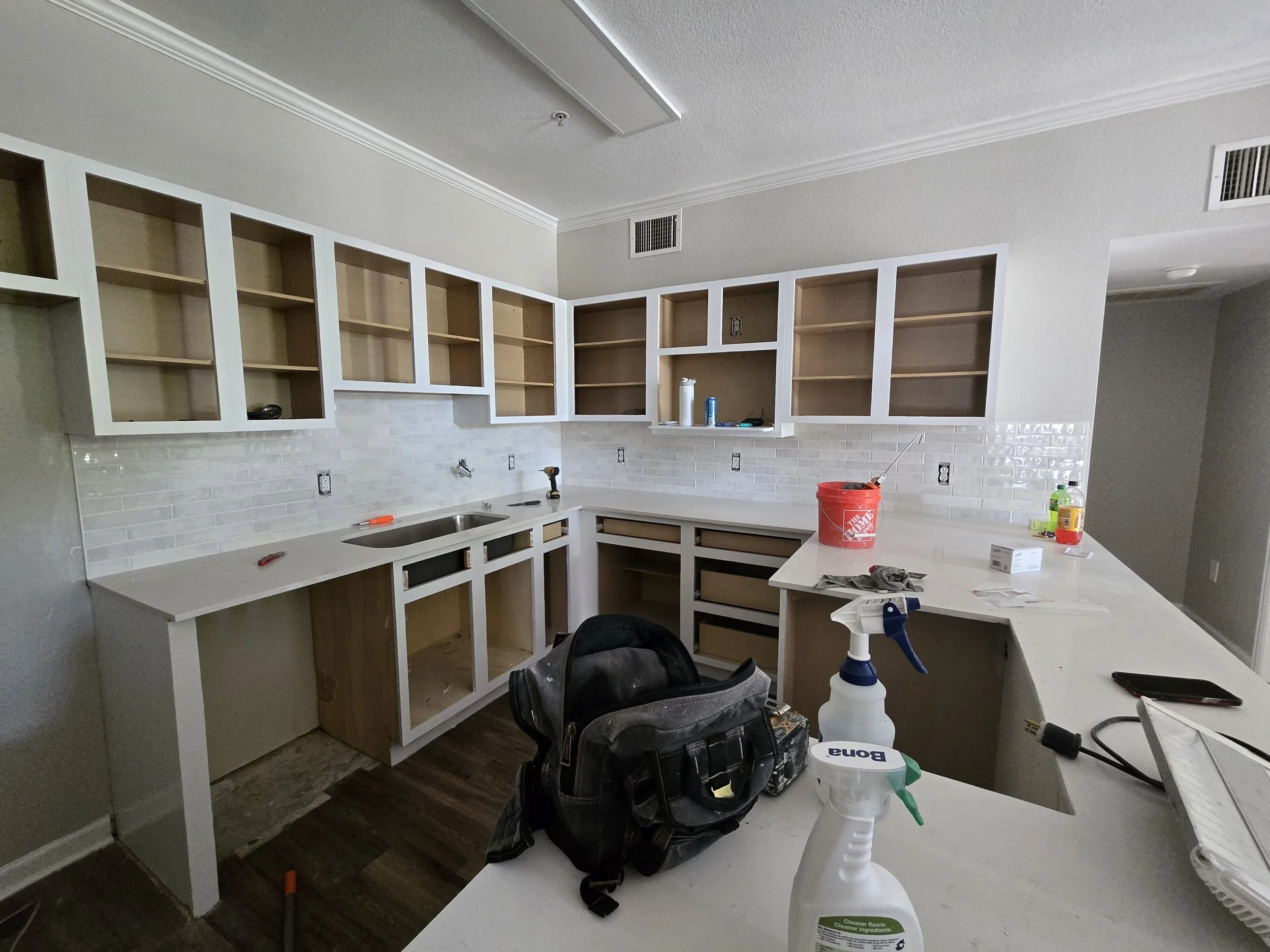 Kitchen under renovation with open cabinets, construction tools, cleaning supplies, a backpack, and a work area with unfinished cabinets and built-in appliances.