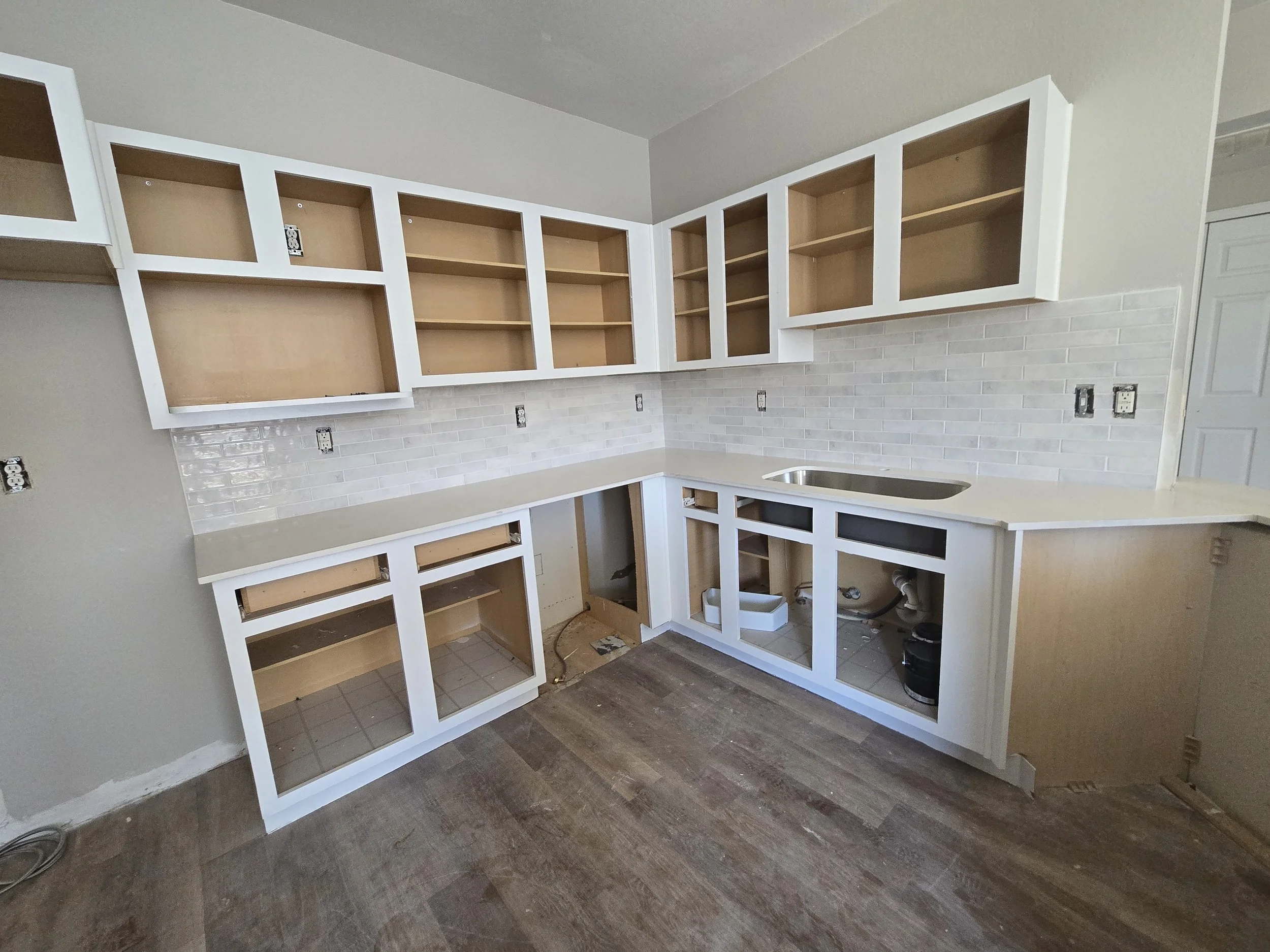Kitchen under renovation with unfinished cabinets, a beige countertop, white brick backsplash, and no appliances installed.