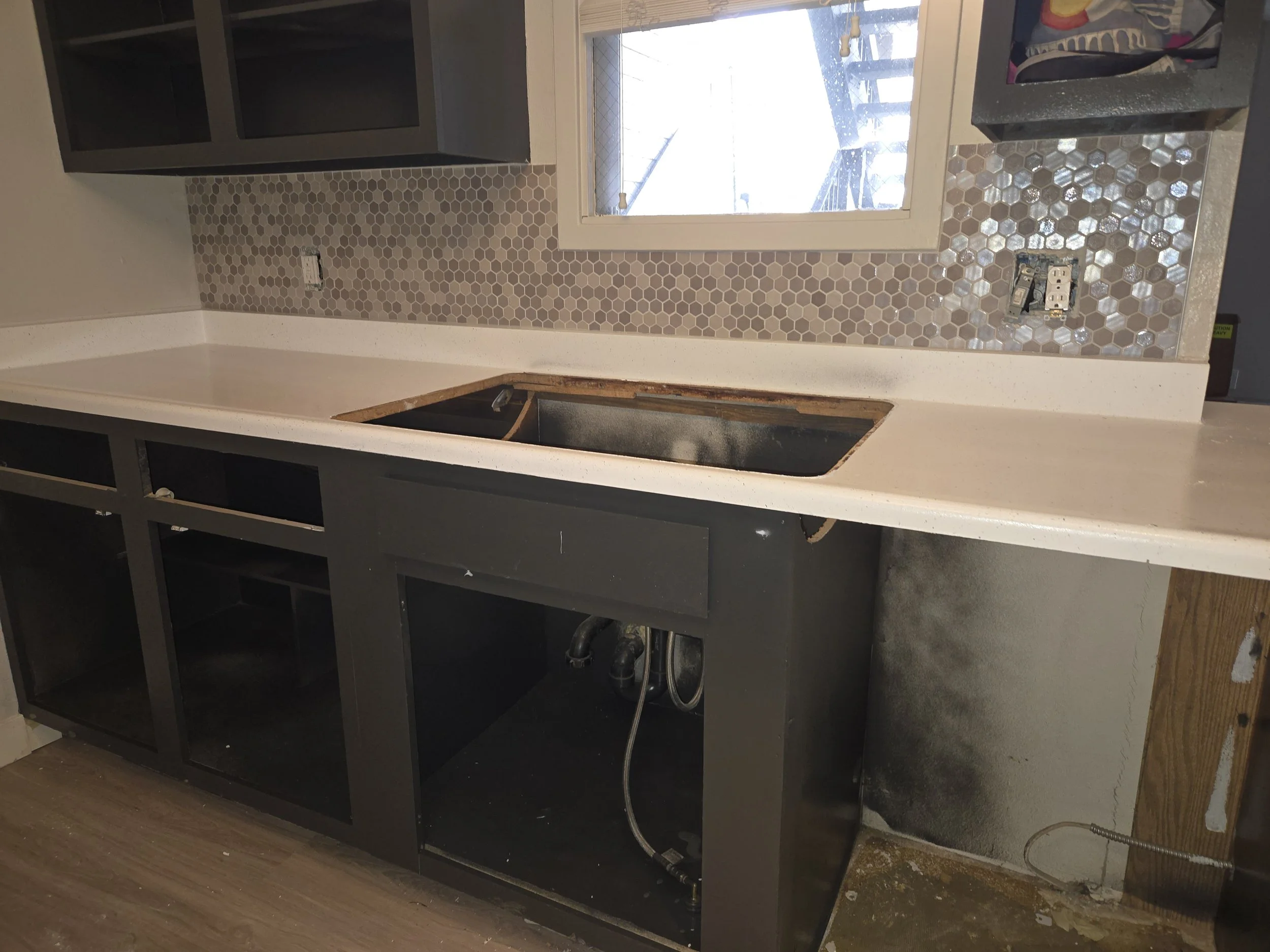 Kitchen cabinet space under construction with a hexagonal tile backsplash, an electrical outlet, and a window. Kitchen sink plumbing visible, black cabinetry, and a countertop. Construction materials and unfinished areas present.