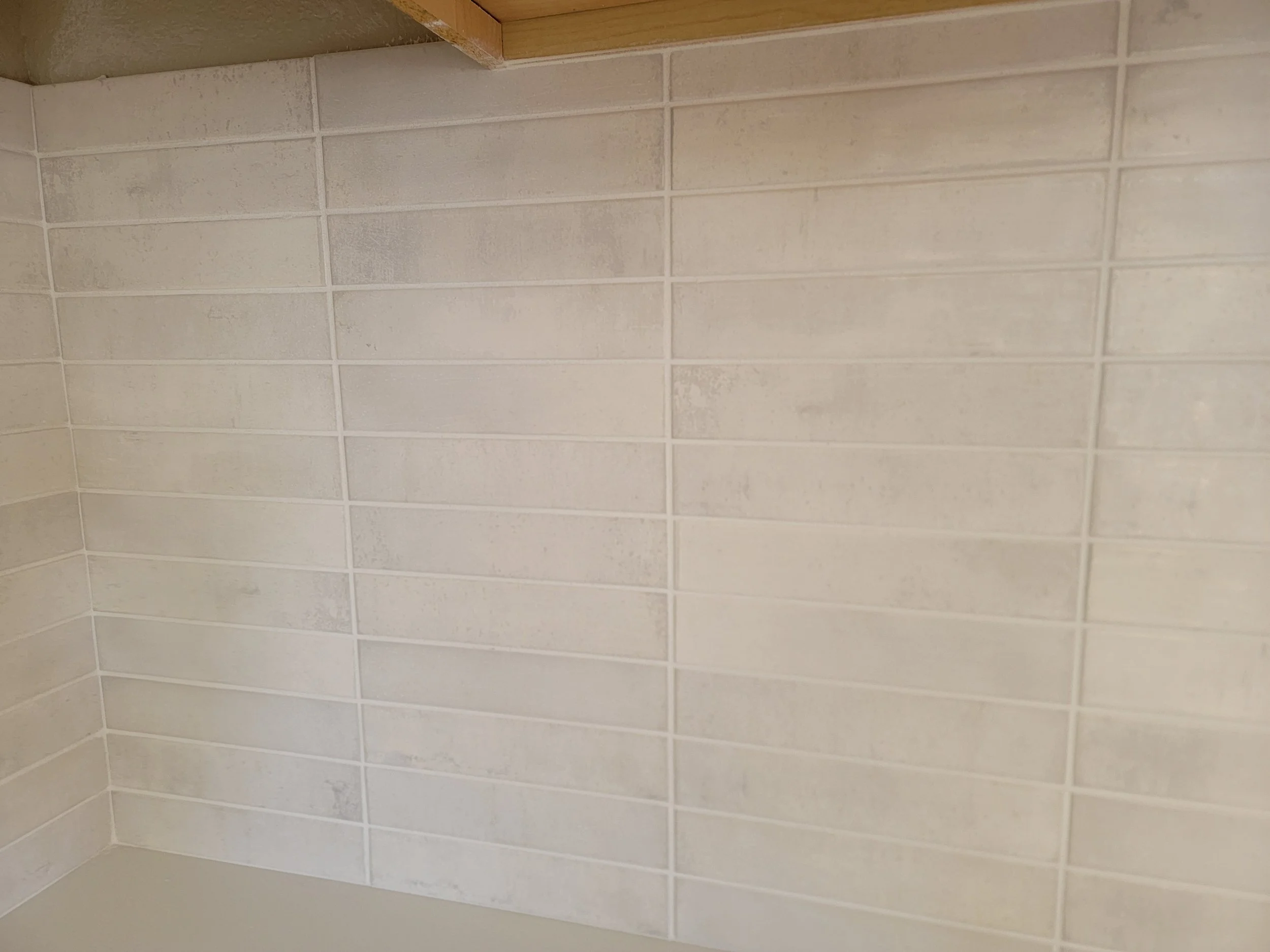 Close-up view of a tiled wall in a bathroom or kitchen, showing beige rectangular tiles with textured surface and white grout lines.