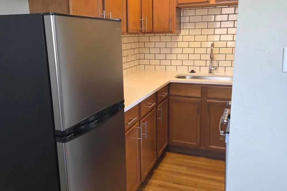 A kitchen with wooden cabinets, a beige countertop, a stainless steel refrigerator, a brick-style backsplash, a sink with a faucet, and partially visible stove on the right.