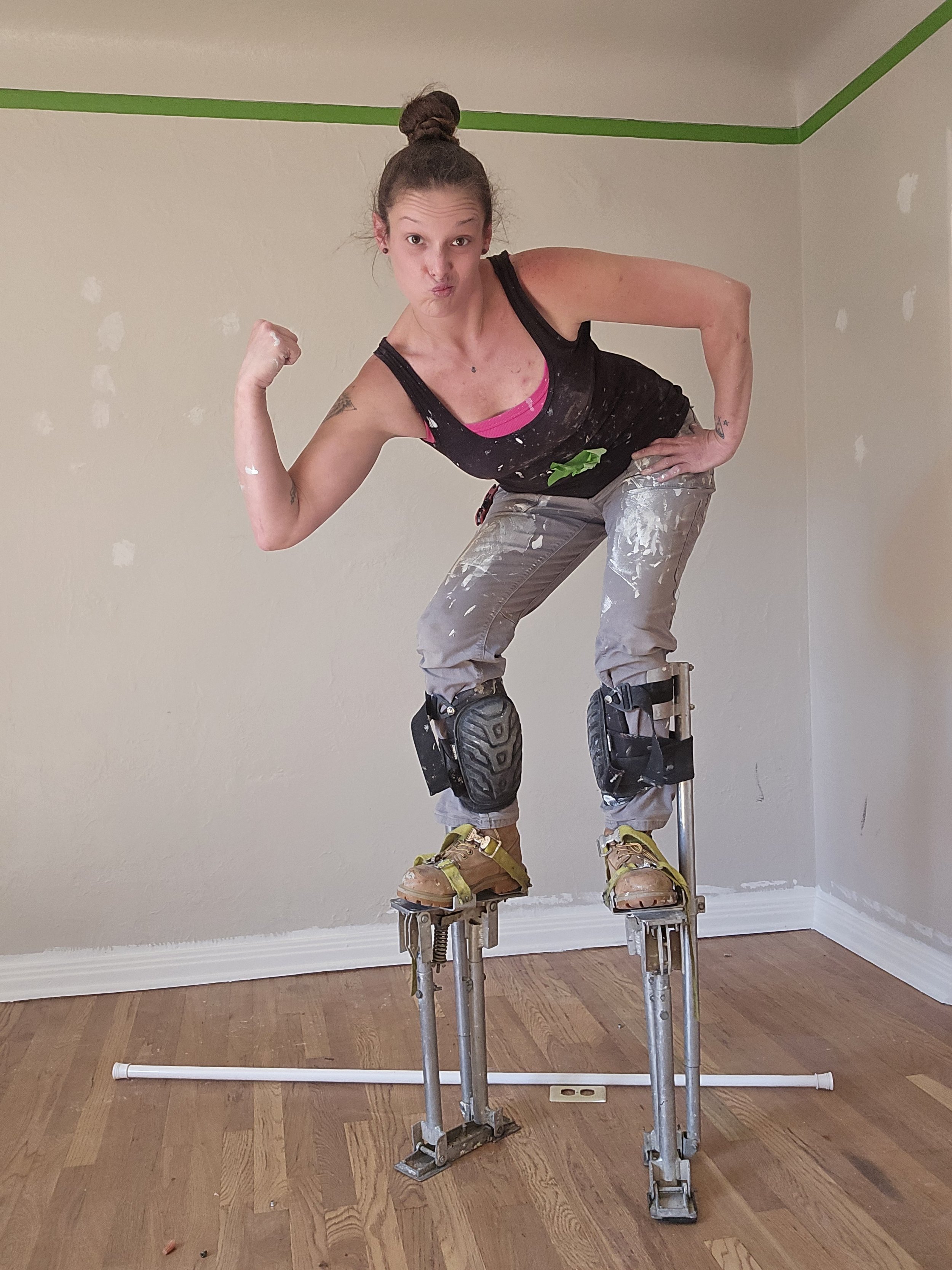 A woman with a rebellious expression, wearing work clothes with paint stains, standing on stilts inside a house with unfinished walls, making a fist with her right hand and resting her left hand on her hip.