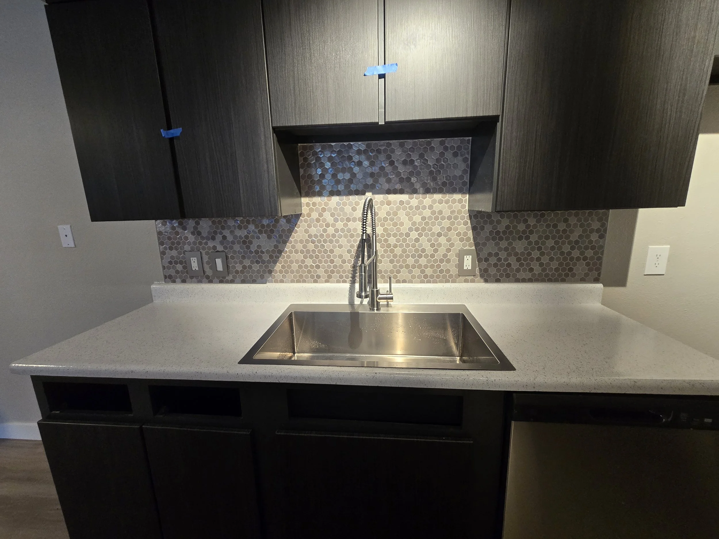 Modern kitchen with dark cabinets, gray quartz countertop, hexagonal tile backsplash, and a stainless steel sink with a high-arc faucet.