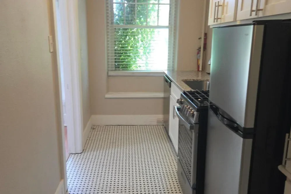 Small kitchen with white cabinets, a window with blinds, a stainless steel refrigerator, a stove, and a countertop with a sink. Tiled black-and-white floor.
