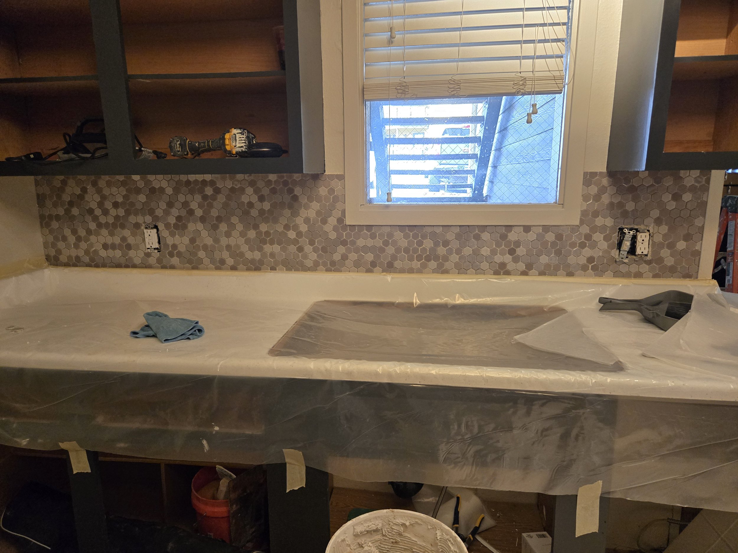 Kitchen with black countertops, hexagonal tile backsplash, a window with blinds, and open shelves. Construction tools and materials are scattered around.