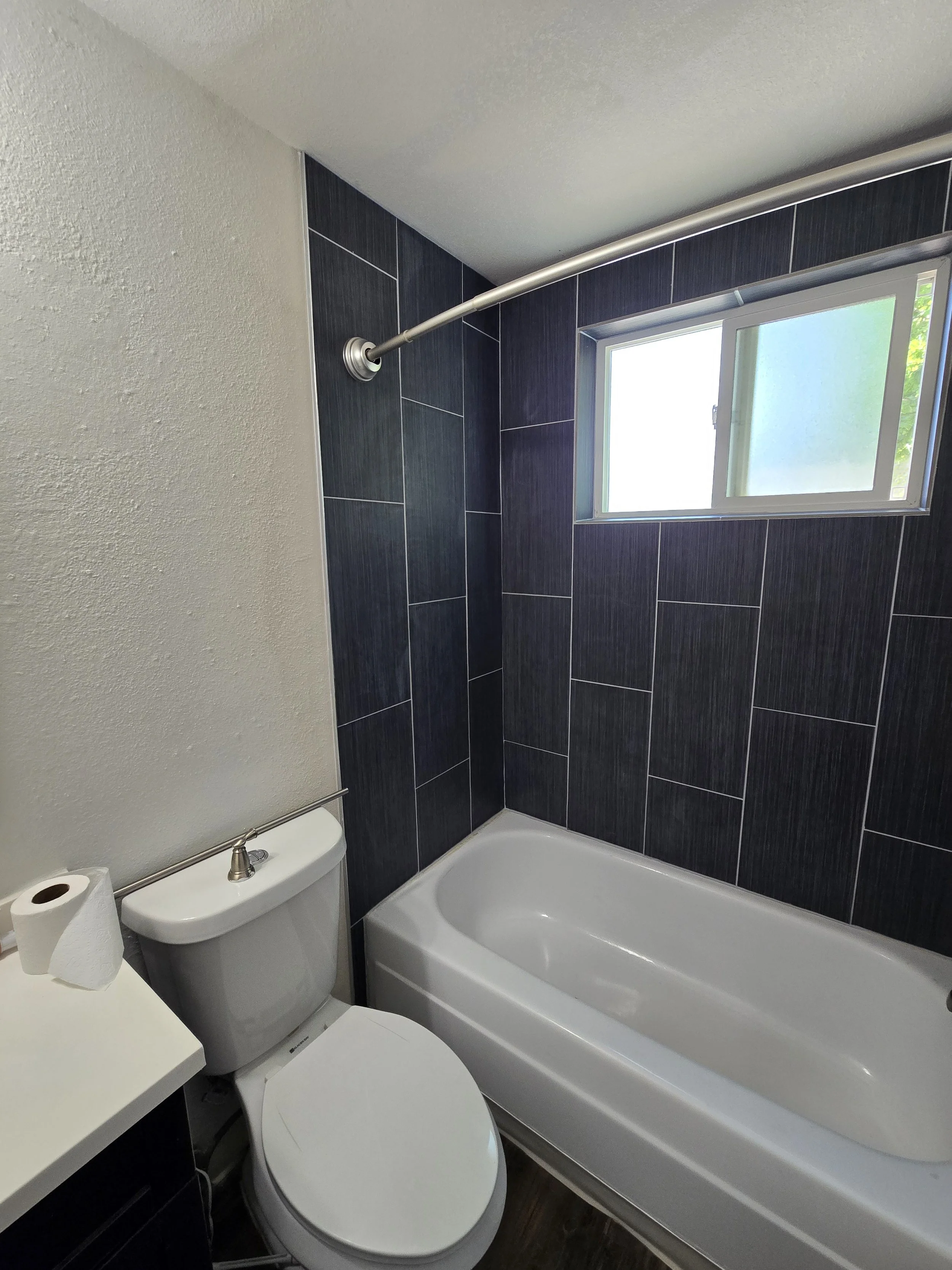 A small bathroom with a toilet, a bathtub, dark tiled wall, a window, and a shower curtain rod.
