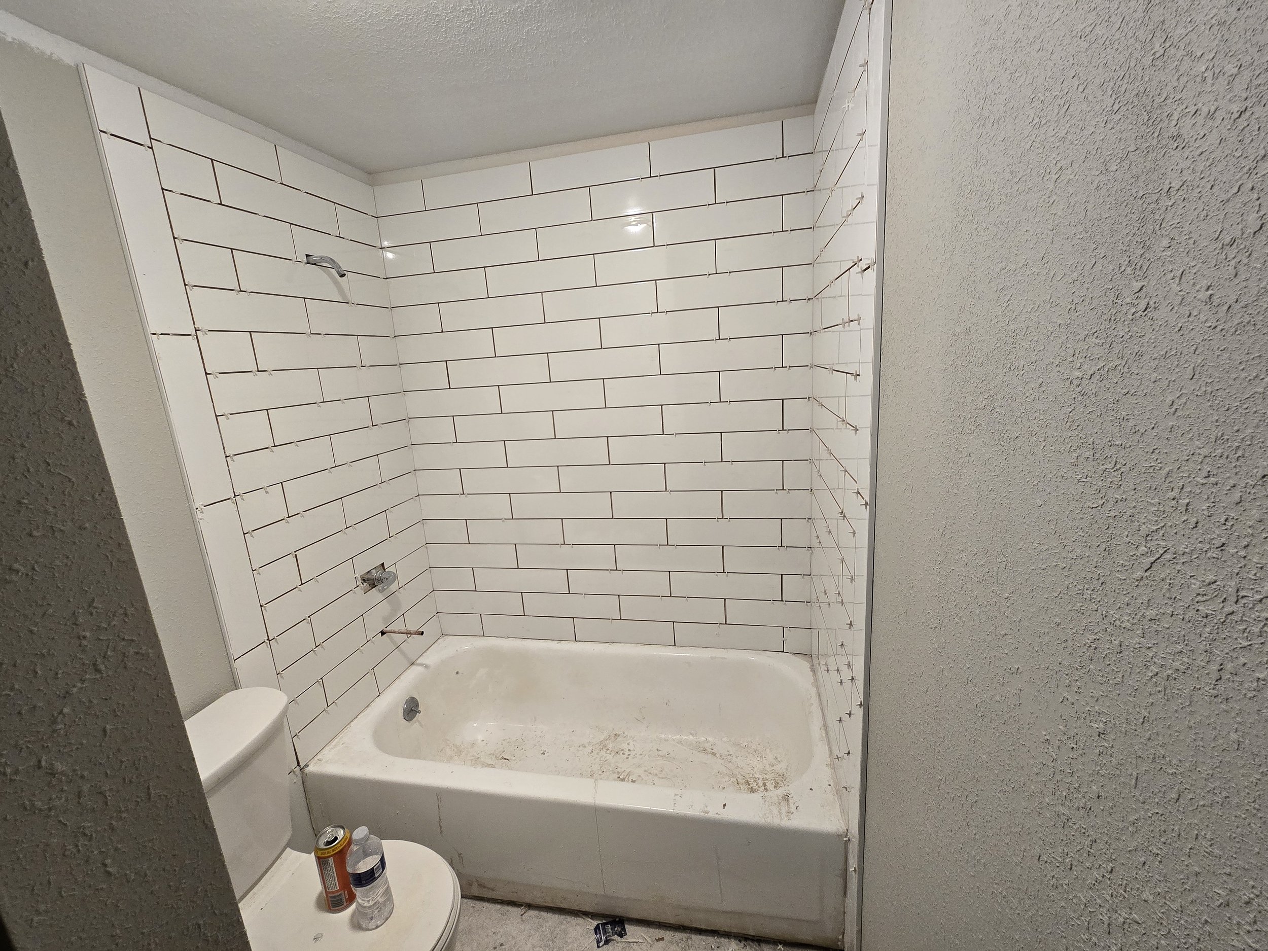 A bathroom under renovation showing a bathtub surrounded by partially installed white subway tiles with brown grout lines, and row of copper pipe connectors along the wall. There are cans of spray paint and a can of soda on the toilet seat in the for
