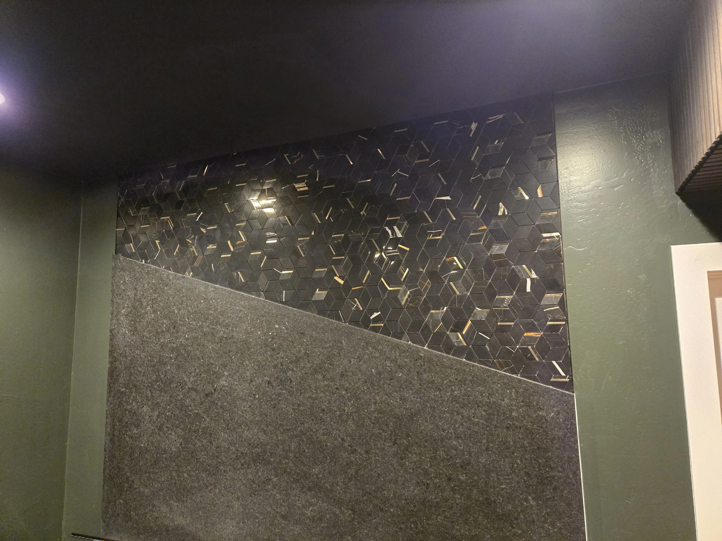 Interior of a room with a decorative wall panel made of black hexagonal tiles with gold accents, green walls, and a speckled black granite countertop.
