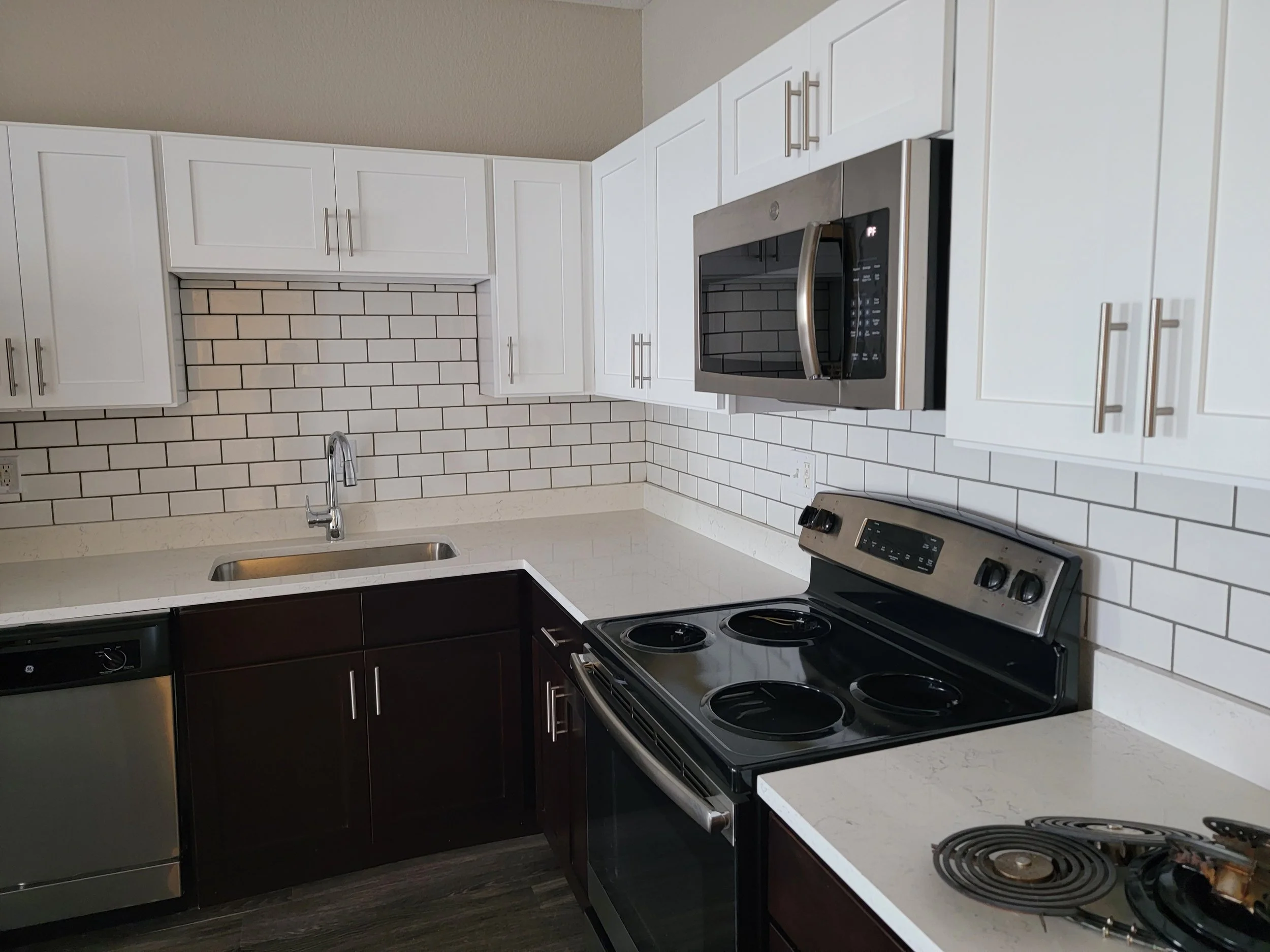 A modern kitchen with white upper cabinets, dark lower cabinets, a white marble countertop, a stainless steel microwave, a stainless steel stove, and a subway tile backsplash. There is a stainless steel sink with a single faucet.