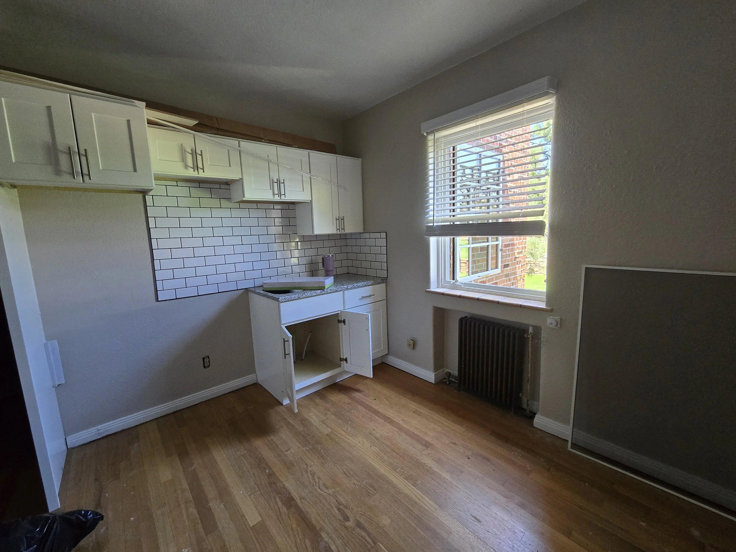 Empty kitchen corner with white cabinets, a window with blinds, a radiator, and a wooden floor.