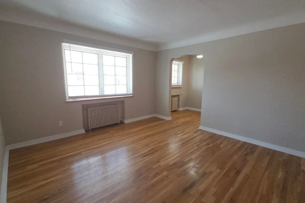 Empty room with hardwood floors, large window with blinds, radiator underneath, and a small alcove with a window and plant.