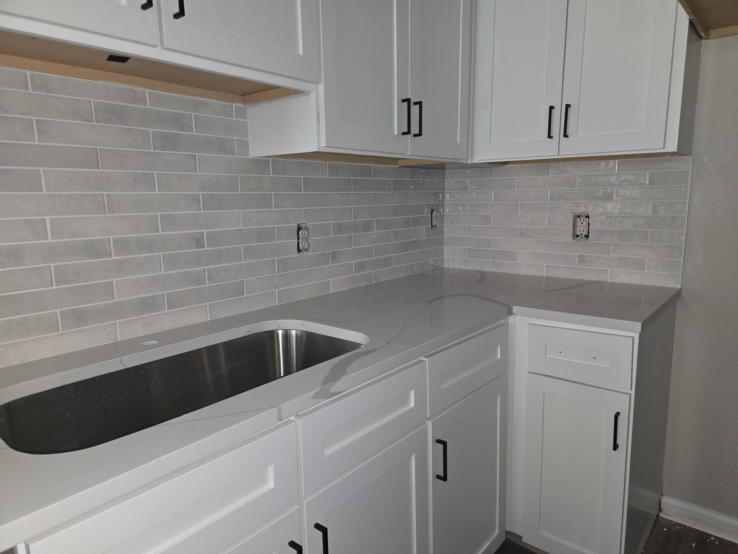 Kitchen with gray tile backsplash, white cabinets with black handles, and a black countertop. Two electrical outlets are visible on the wall.