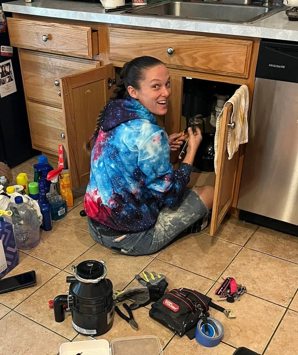 Woman wearing a galaxy print hoodie kneeling on kitchen floor, fixing an under-sink garbage disposal, with tools and cleaning supplies around her.