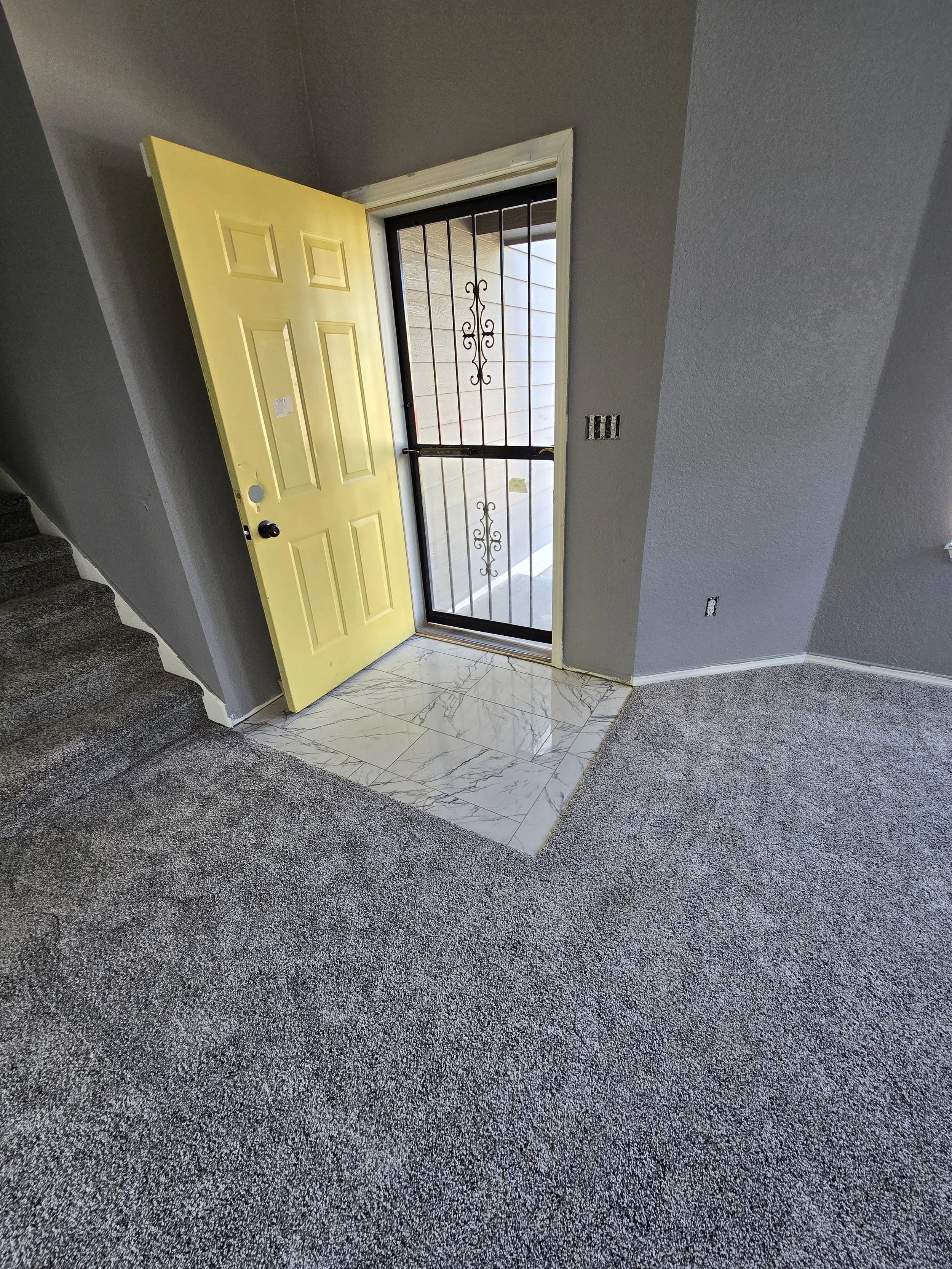 Open yellow front door with a black door knob and a security grill, leading to a porch with white wood siding and marble tiled entryway, interior with gray walls and plush gray carpet.