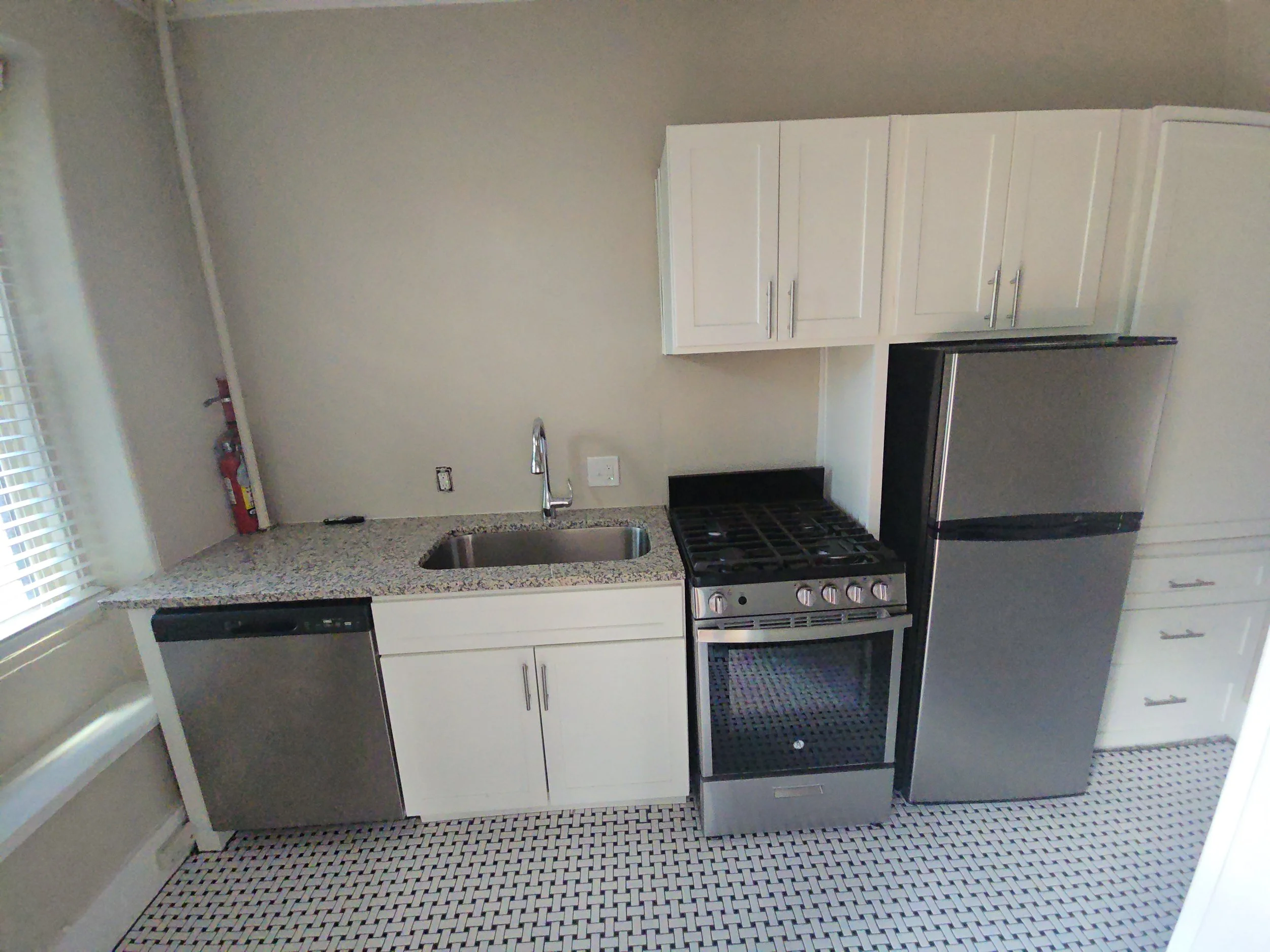 Modern kitchen with white cabinets, stainless steel oven, and black dishwasher. Granite countertop, black and white patterned tile floor, window with blinds, and beige wall.