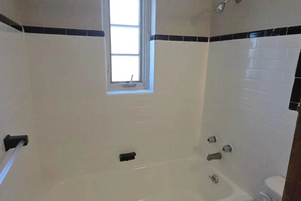 Bathroom with white tiled walls, black trim, a window, and fixtures for a bathtub or shower.