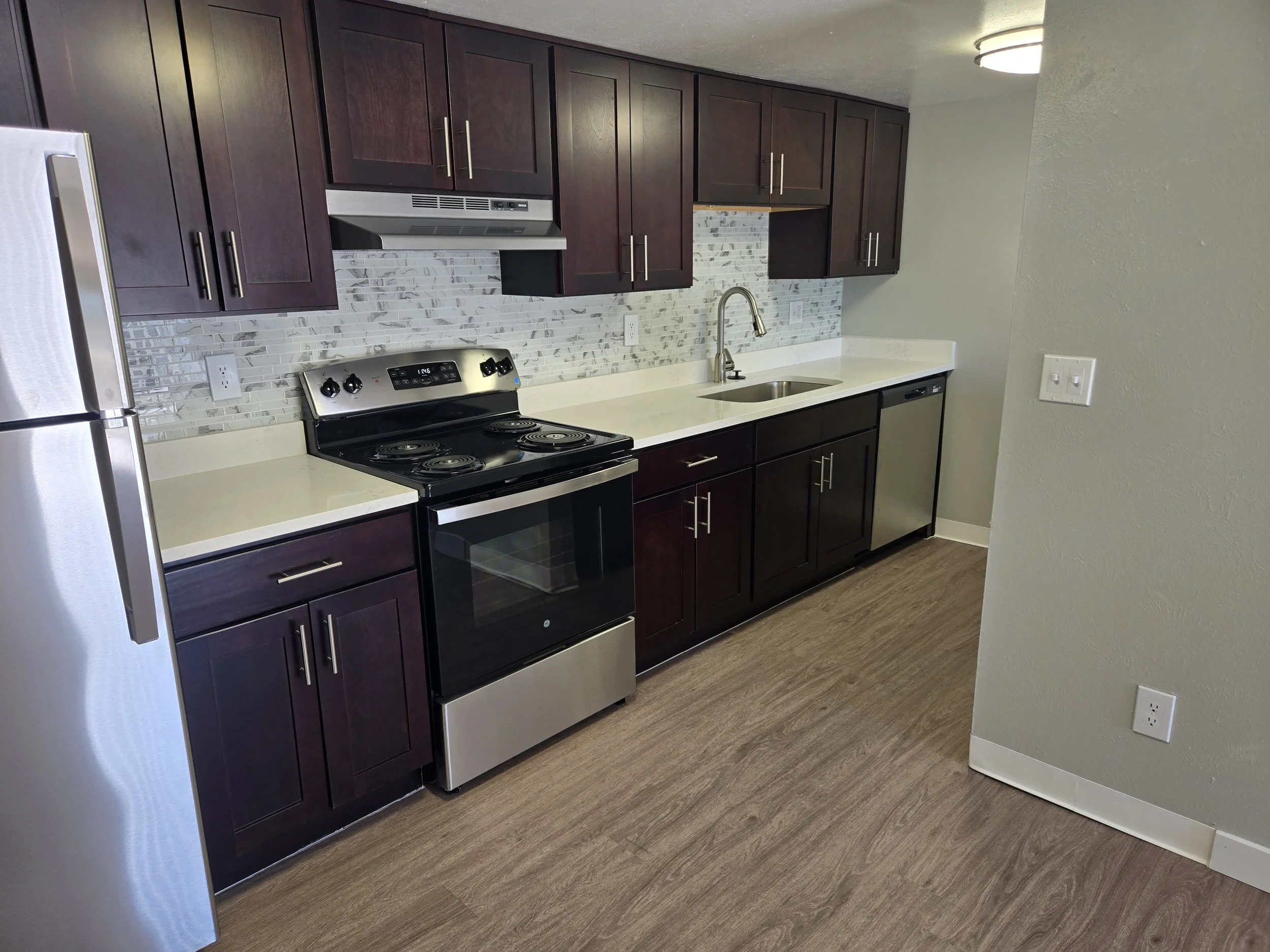 Modern kitchen with dark wood cabinets, stainless steel appliances including refrigerator and oven, marble backsplash, and beige countertop with a stainless steel sink.