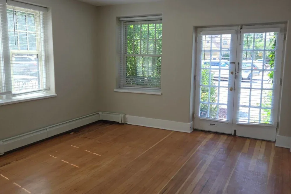 Empty room with hardwood floors, two windows, and a glass door with blinds, overlooking a parking lot and greenery outside.