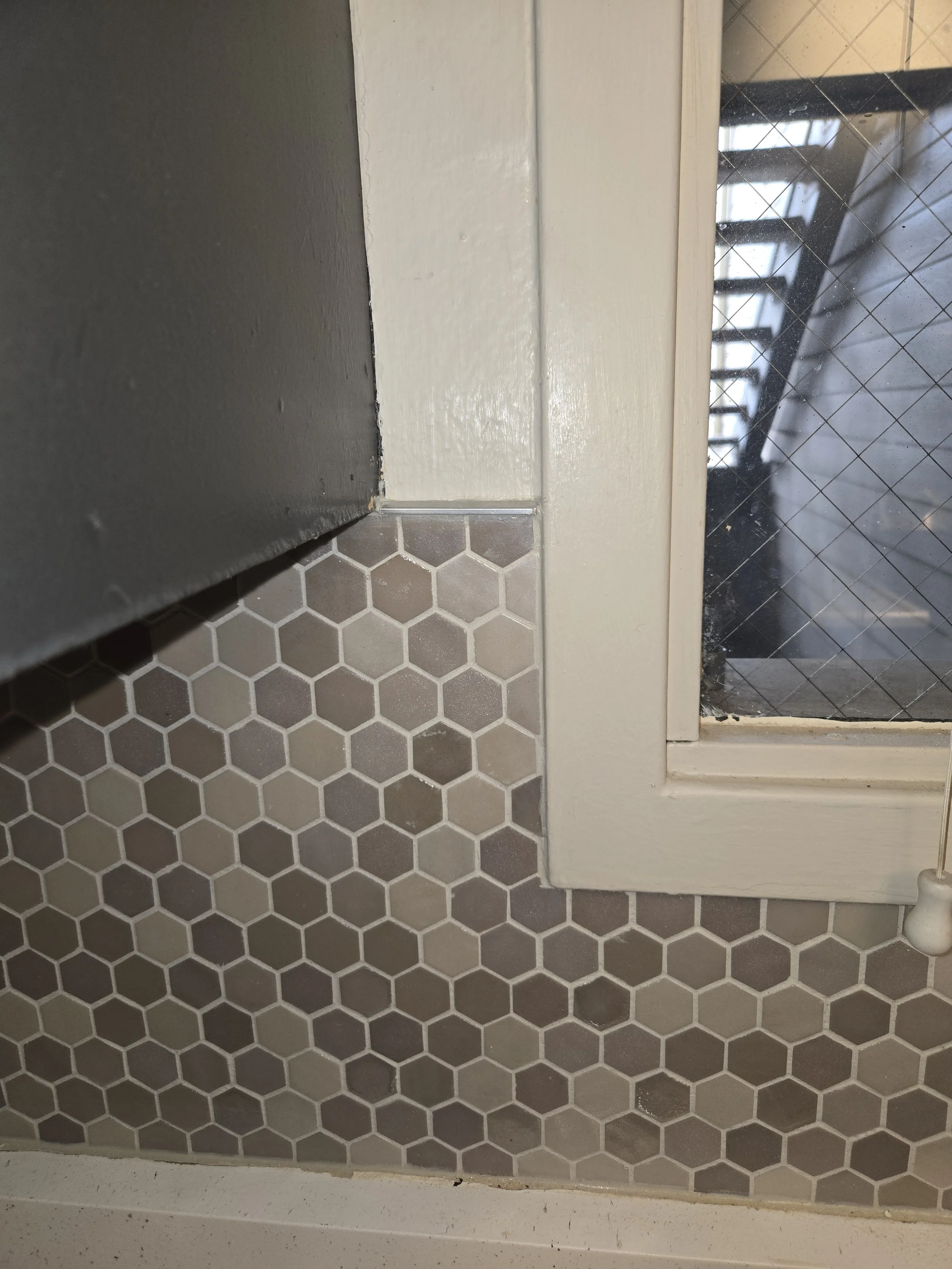 Close-up view of a kitchen or bathroom window with a white frame, a black wire mesh screen, and beige tile flooring with a hexagonal pattern.