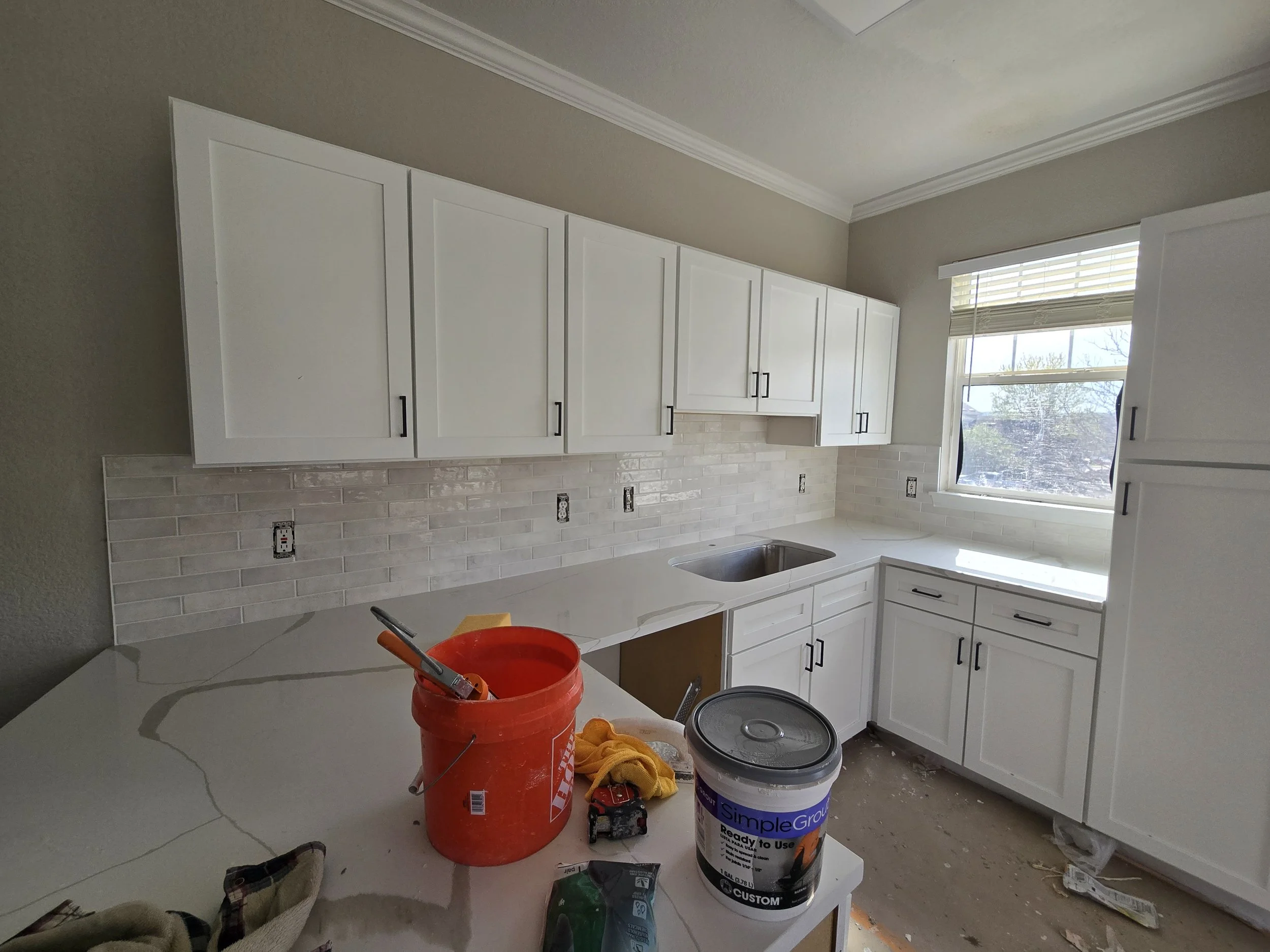 Kitchen under renovation with white cabinets, part of the wall tiled, and construction supplies including a red bucket, paint cans, and tools on the counter.