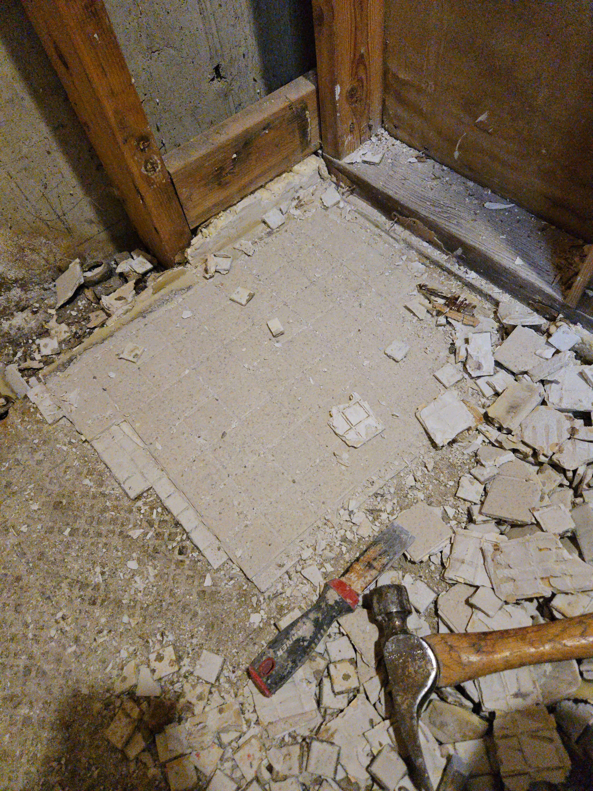 Construction site with tiled floor in the process of removal or installation. Several broken tiles and debris scattered around. A carpenter's square, a hammer, and a power drill are on the floor. There are wooden framing and a wall in the background.