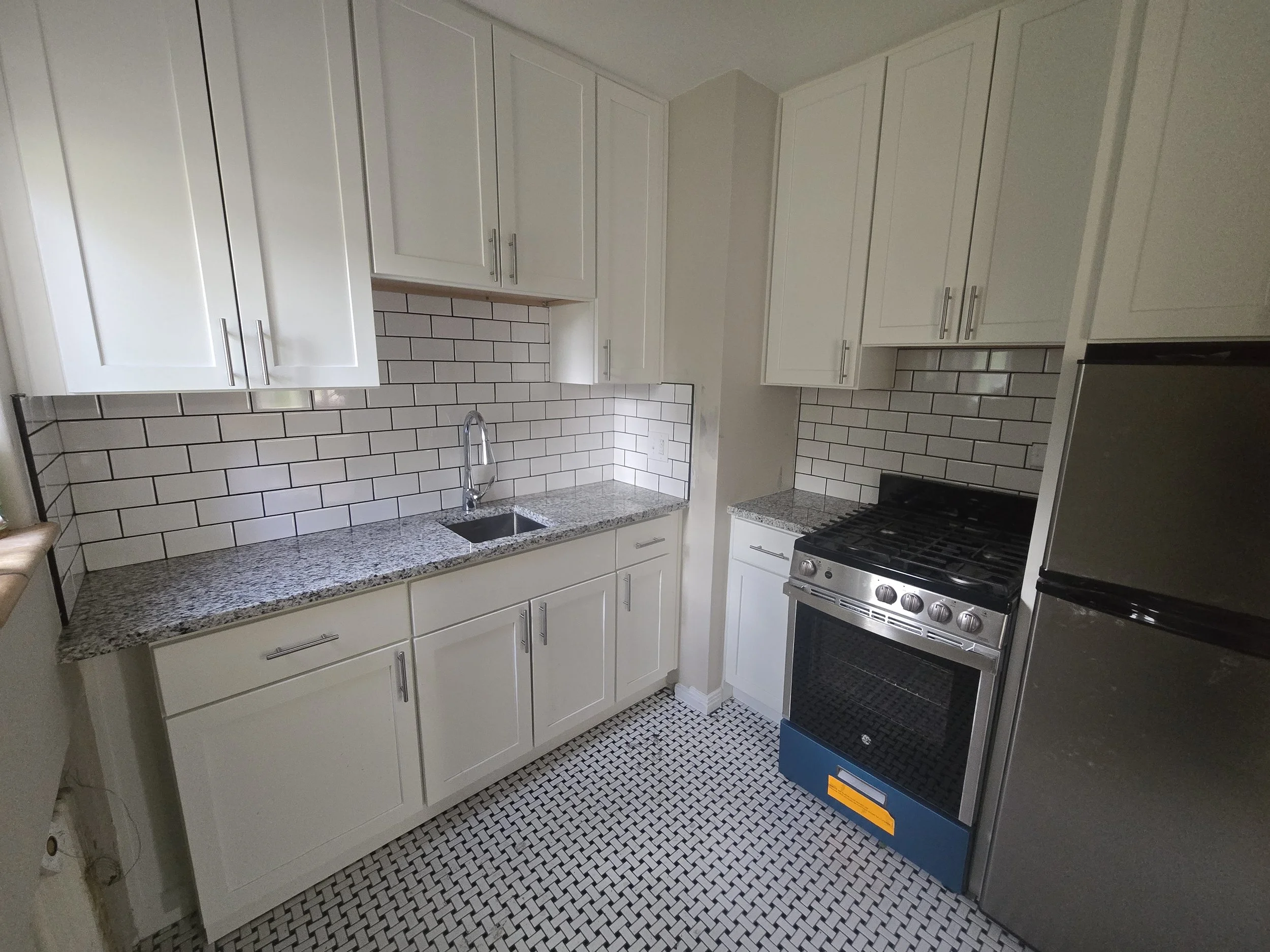 A modern kitchen with white cabinets, subway tile backsplash, granite countertops, a stainless steel sink, and a stove with an oven. The floor has small black and white tile patterns.