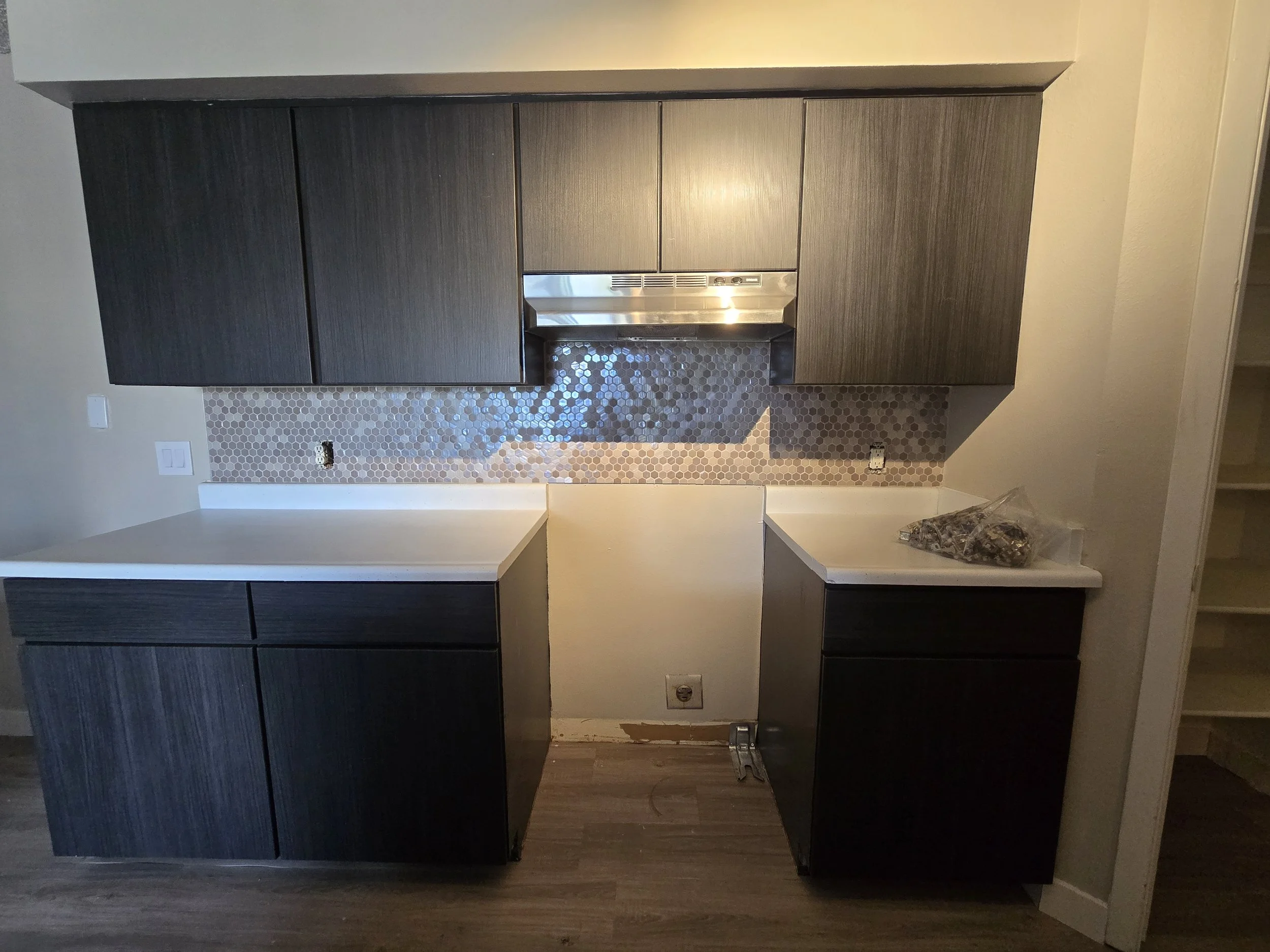 Kitchen space with dark gray cabinets, a range hood, and a hexagonal tile backsplash, under construction or renovation with exposed outlets and unfinished edges.