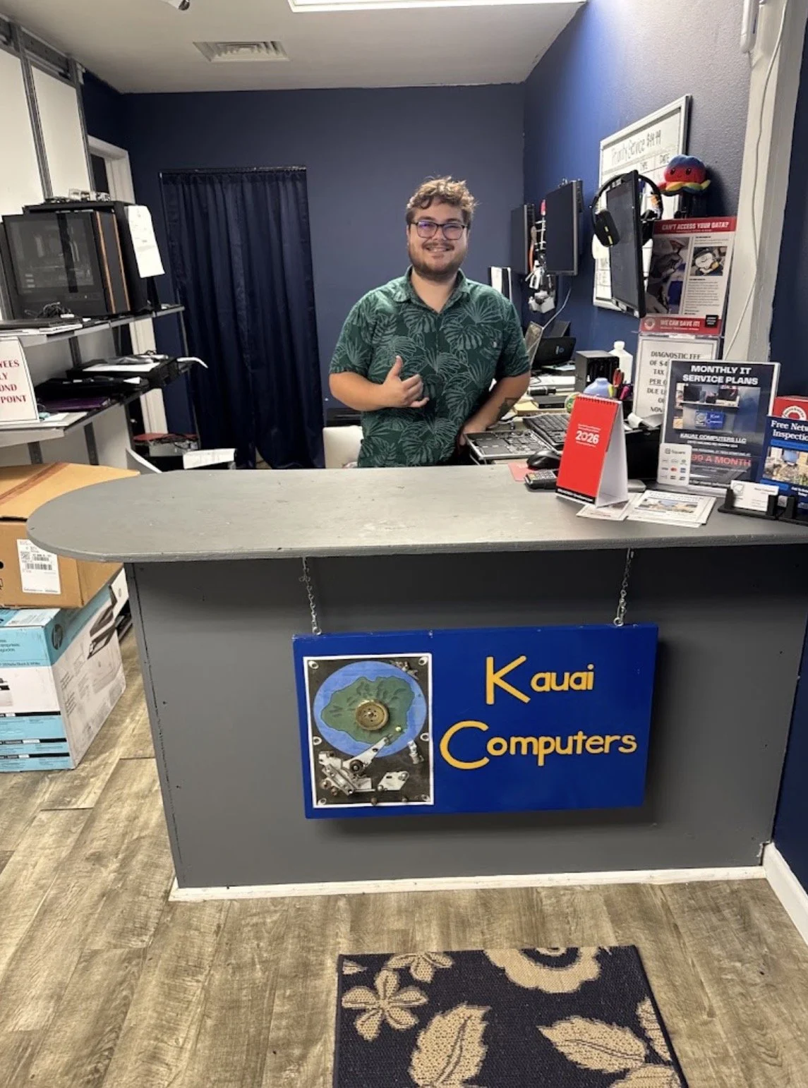 A smiling man with glasses behind a desk at Kauai Computers, a small electronics store. The desk has a sign with the store's name and an image of a computer motherboard. The store has shelves with electronic devices and posters on the wall.