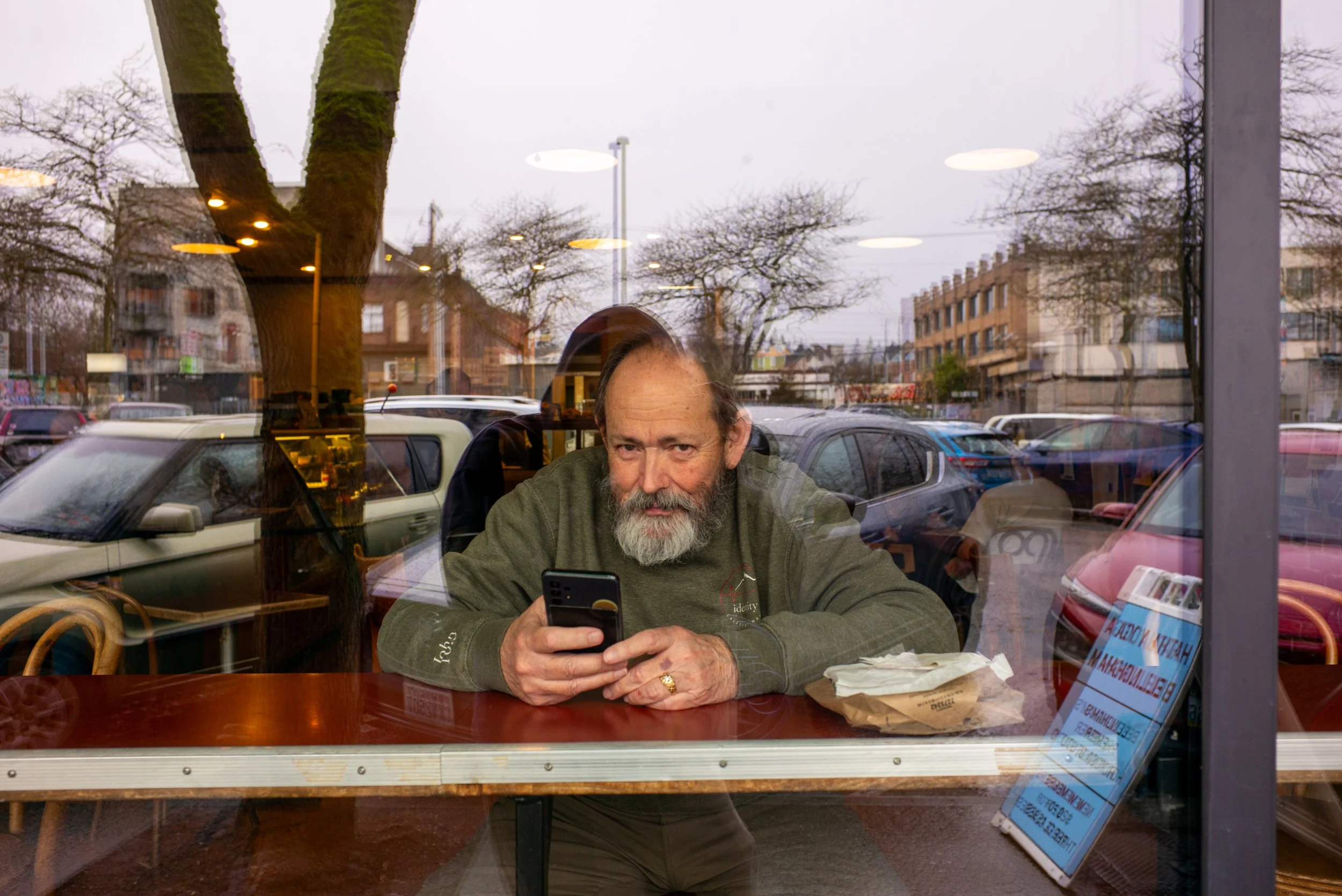 A bearded man sitting at a table inside a cafe, looking at his smartphone, with a large window behind him reflecting parked cars, trees, and buildings outside, and a few illuminated ceiling lights visible through the window.