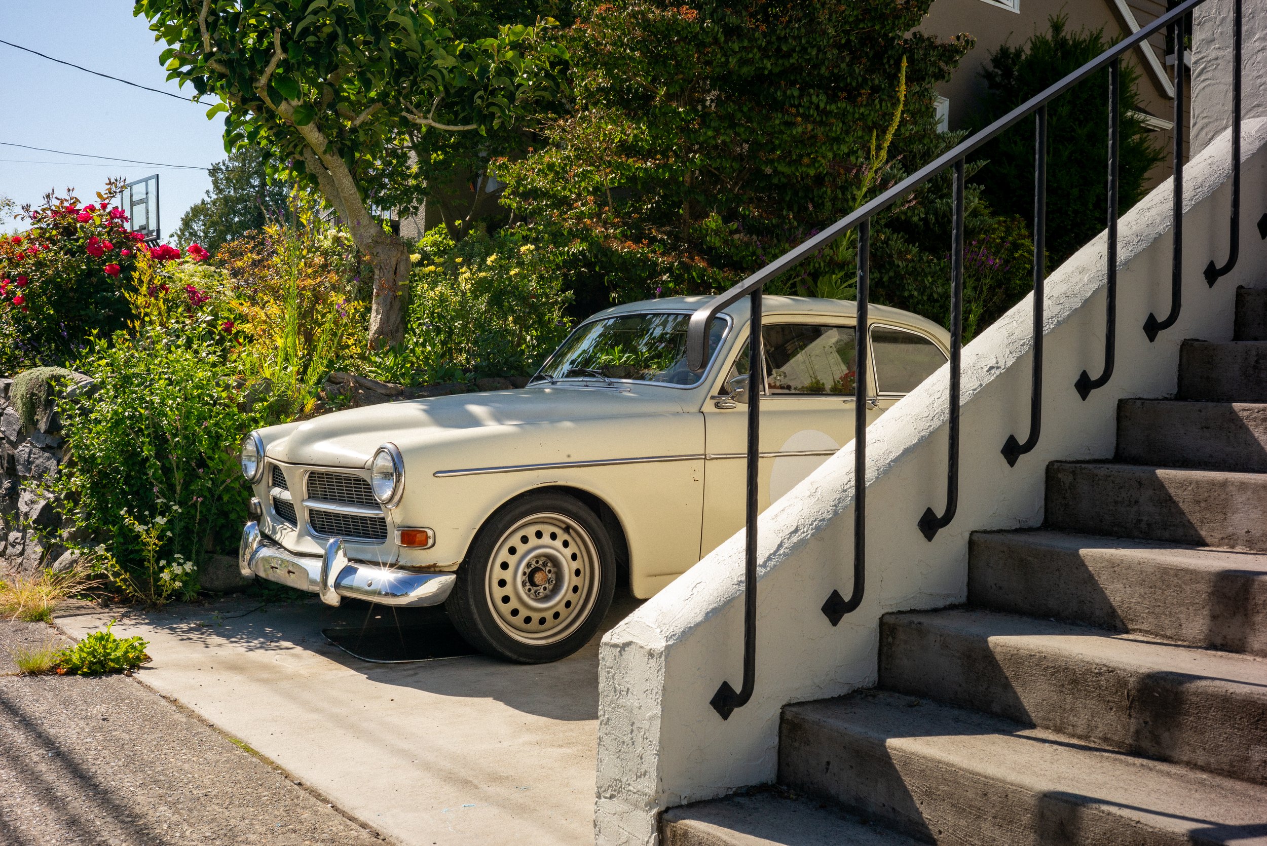 A vintage white car parked under stairs leading to a building, with lush green trees and colorful flowers in the background.
