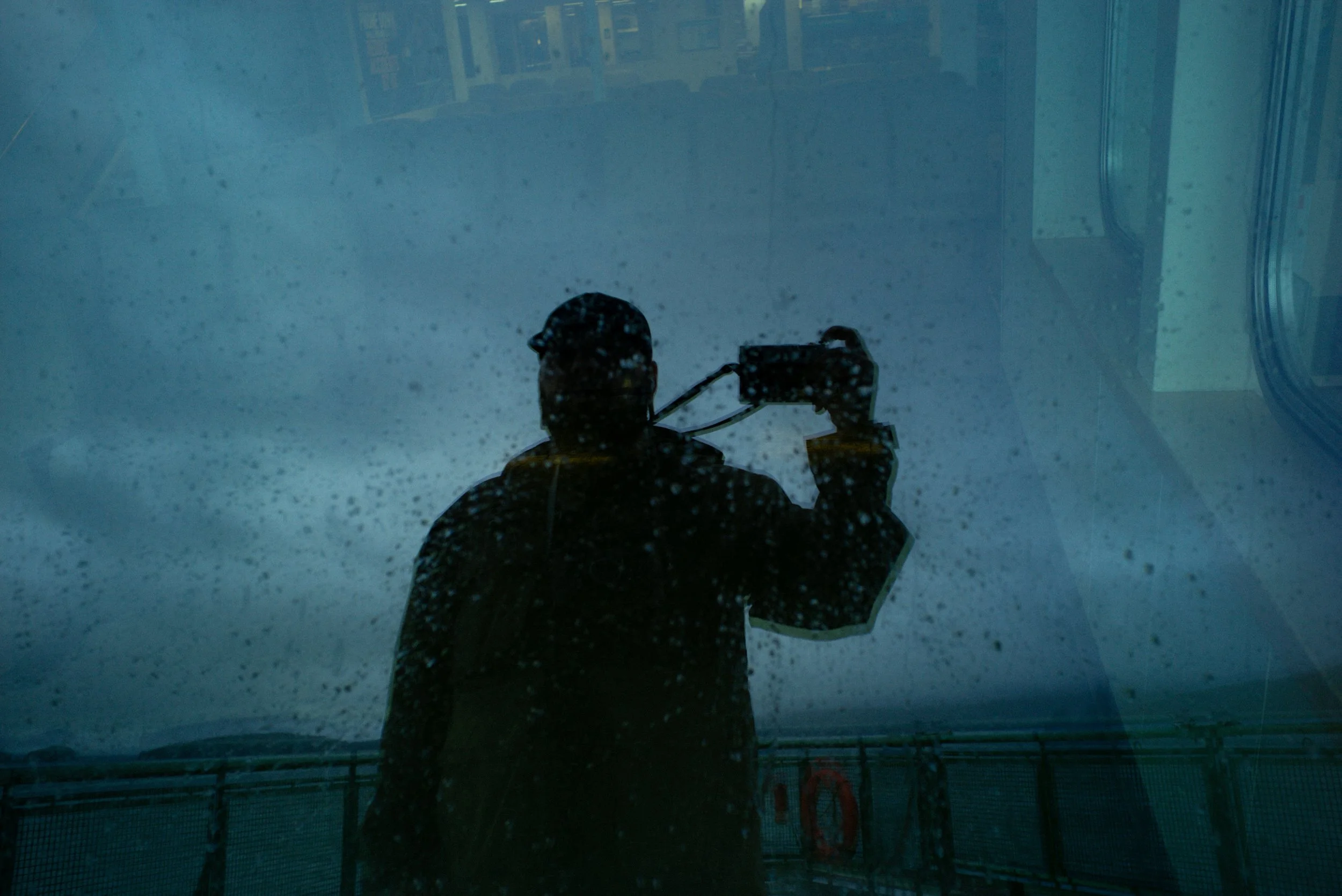 A person taking a photo of their reflection on a rain-covered glass window or surface, with a background featuring a cloudy sky and a railing.