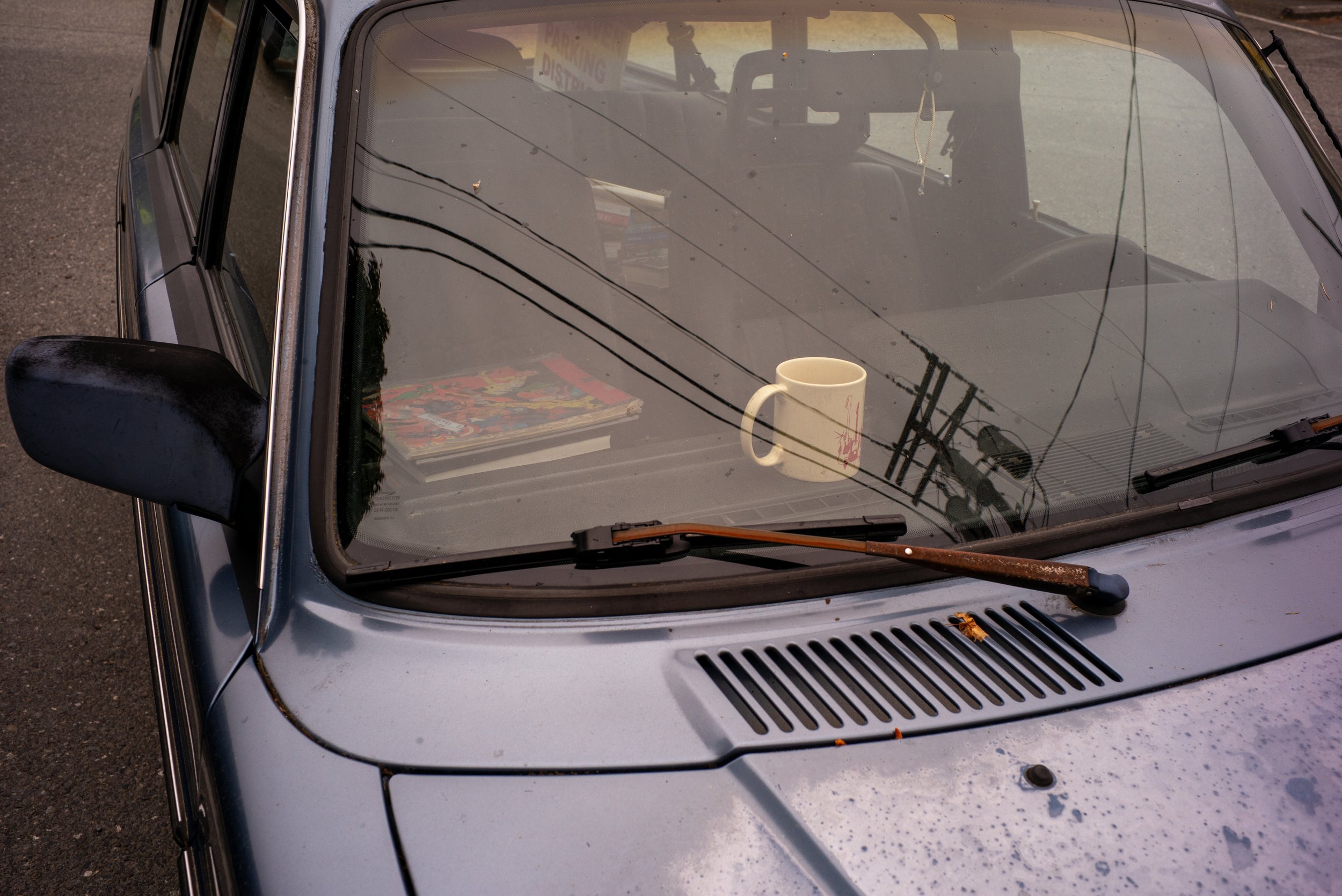 Front view of an old gray car with a white mug on the dashboard, a magazine, and reflections on the windshield.