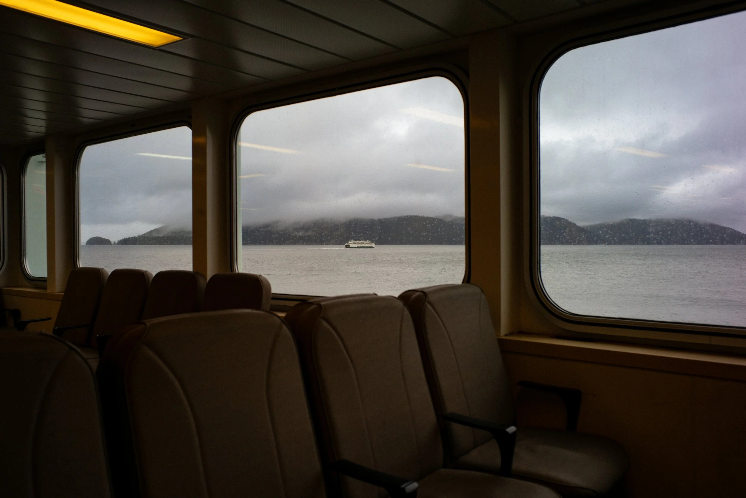 Inside view of a boat or ferry with empty seats, looking out large windows at cloudy weather and an island with a boat in the water.