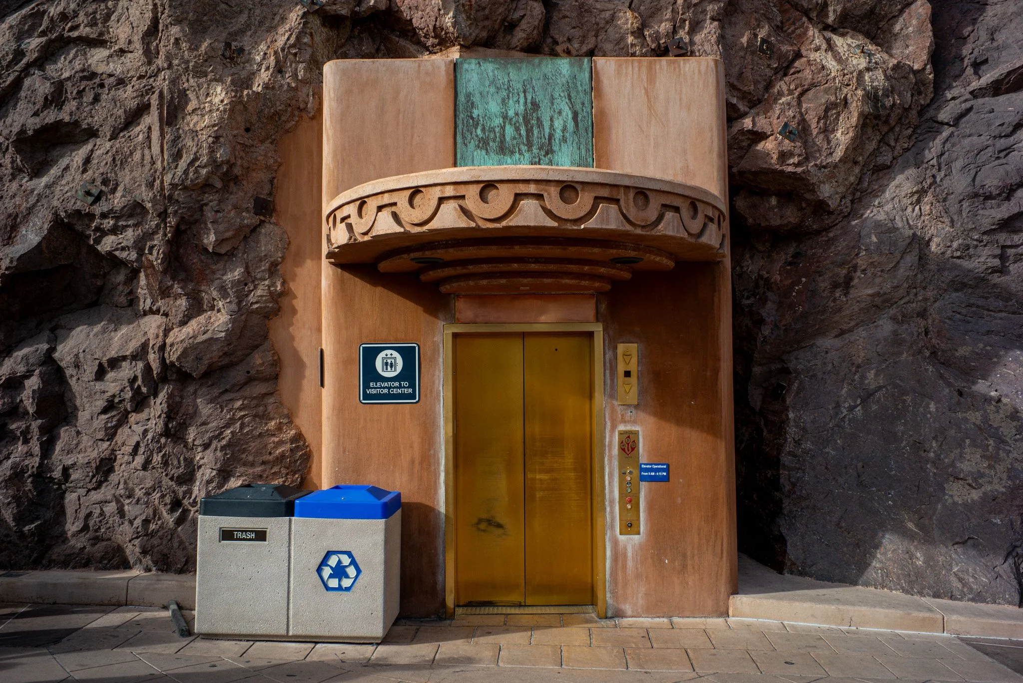 Elevator doors set into a rocky wall with a blue sign indicating elevator operation hours, a trash bin labeled 'TRASH' with a recycling symbol, and a recycling bin in front.
