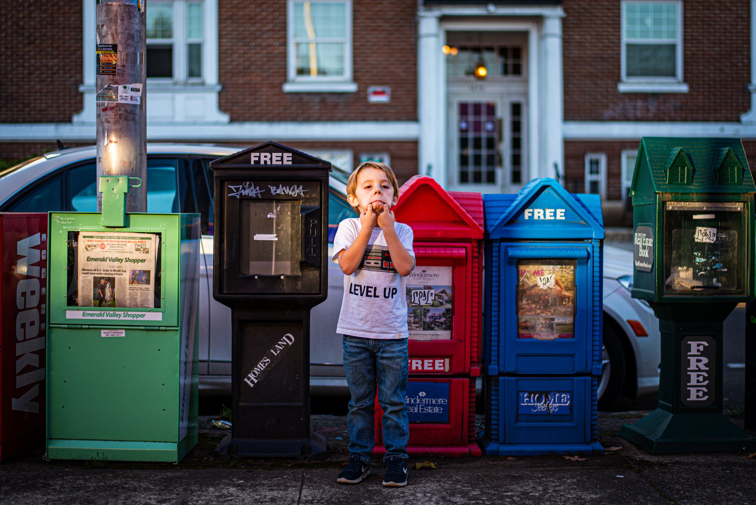 A young boy standing in front of a row of colorful newspaper vending machines on the sidewalk, with a brick building and windows in the background, during what appears to be early evening.