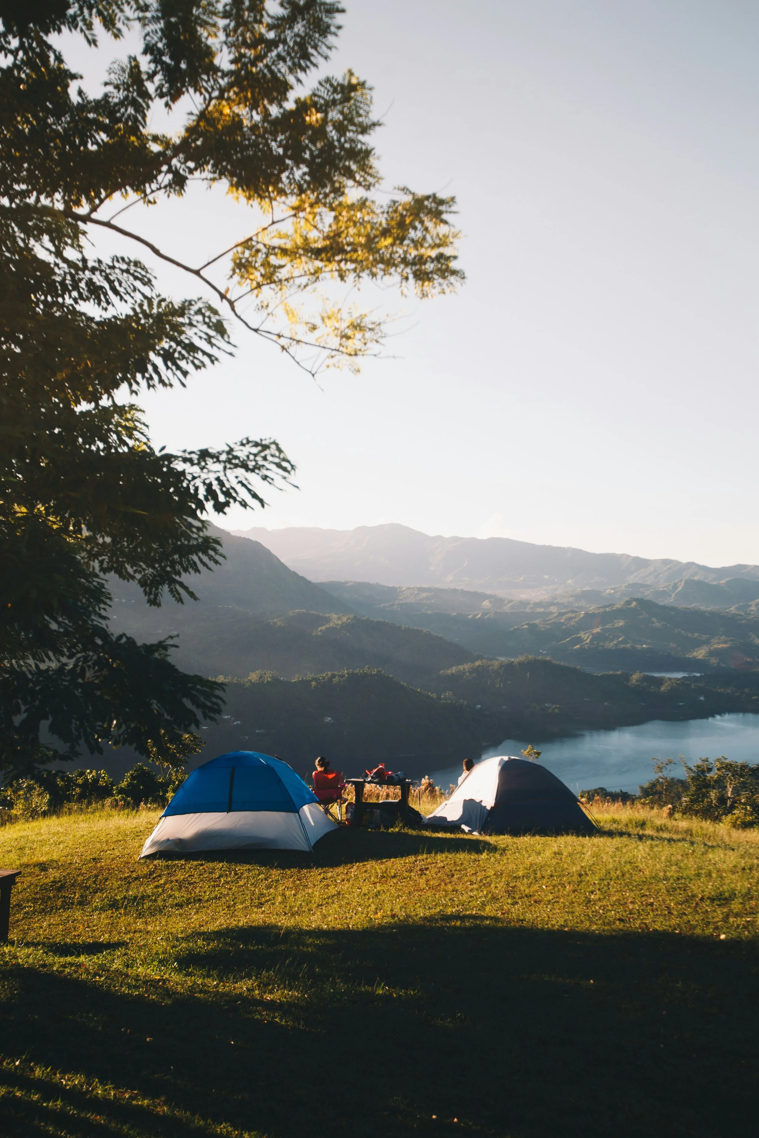 A scenic outdoor camping scene with three tents on a grassy area near a lake, surrounded by mountains and trees, with campers relaxing and enjoying the view during sunset.