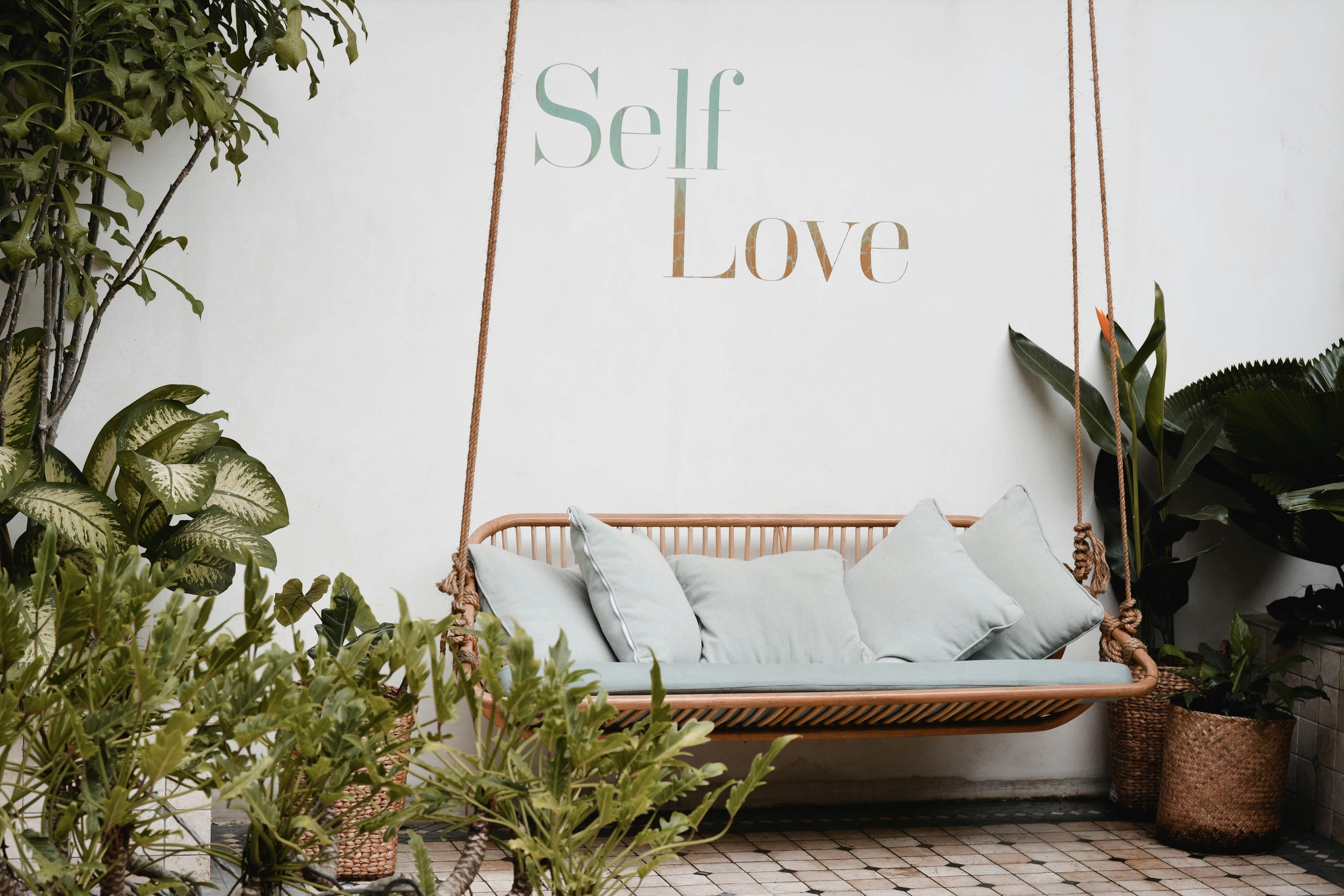 A hanging wooden swing with white cushions in front of a white wall decorated with a sign that says 'Self Love'. Green plants surround the swing, creating a cozy, natural atmosphere.