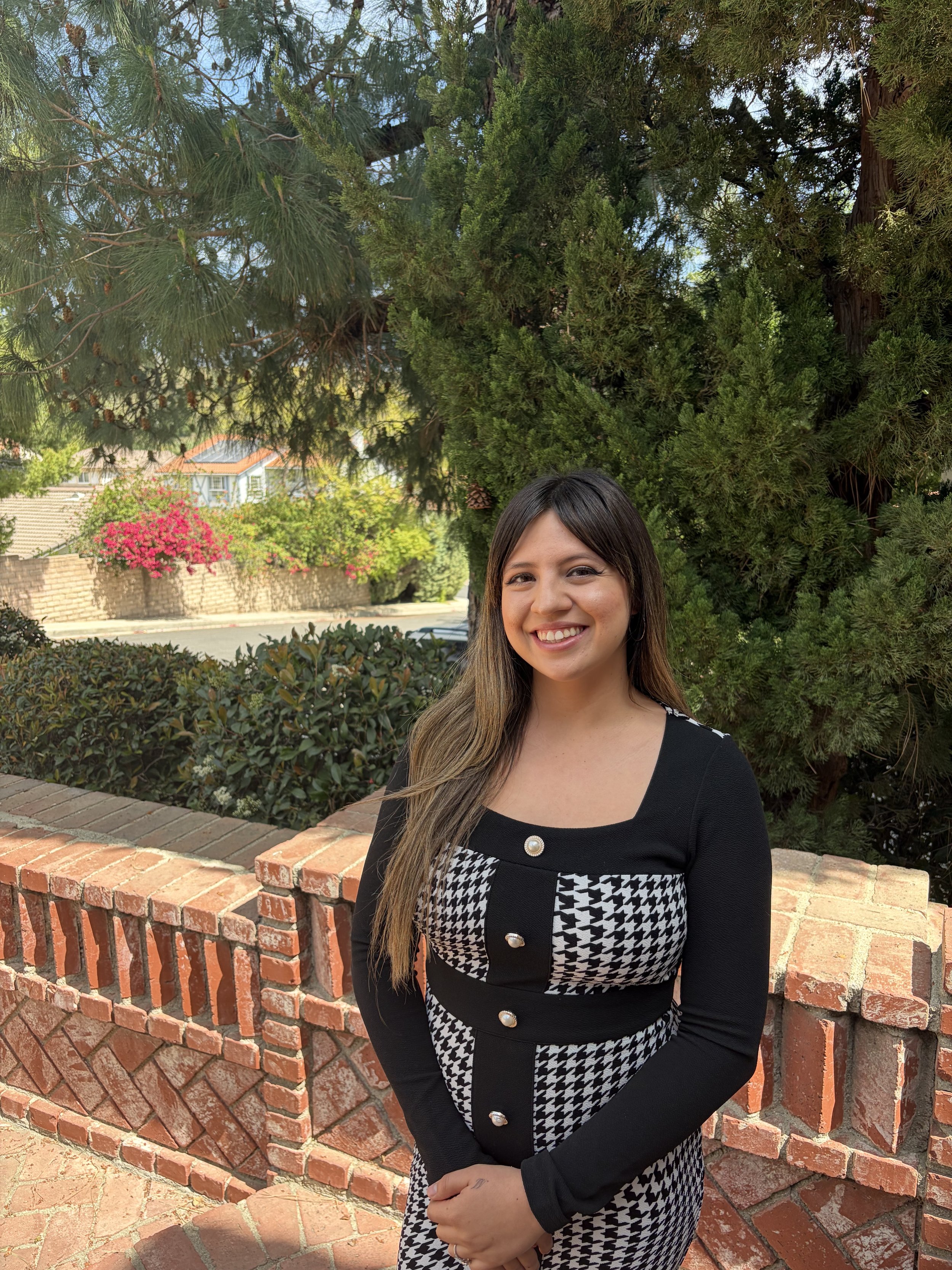 A woman with long brown hair smiling outdoors near a brick wall, with large green trees in the background.