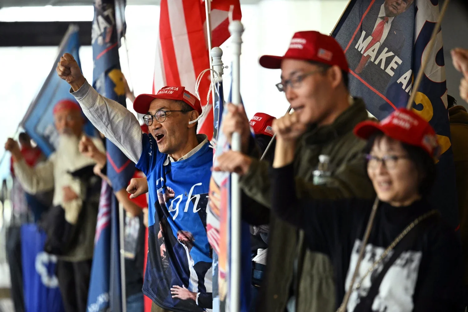 Group of supporters at a rally, many wearing red 'Make America Great Again' hats, holding flags, and cheering passionately.