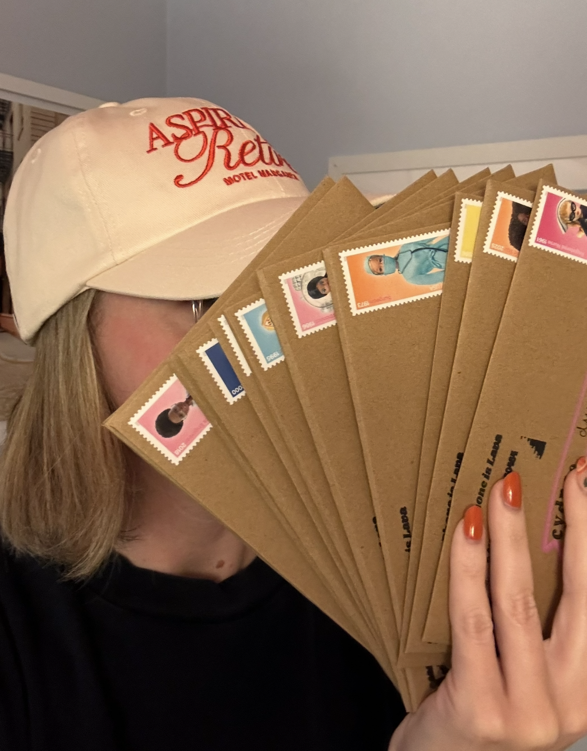Person wearing a beige cap with red embroidered text 'Aspire Rest Motel & Lounge' and holding multiple brown envelopes each decorated with colorful postage stamps and images.