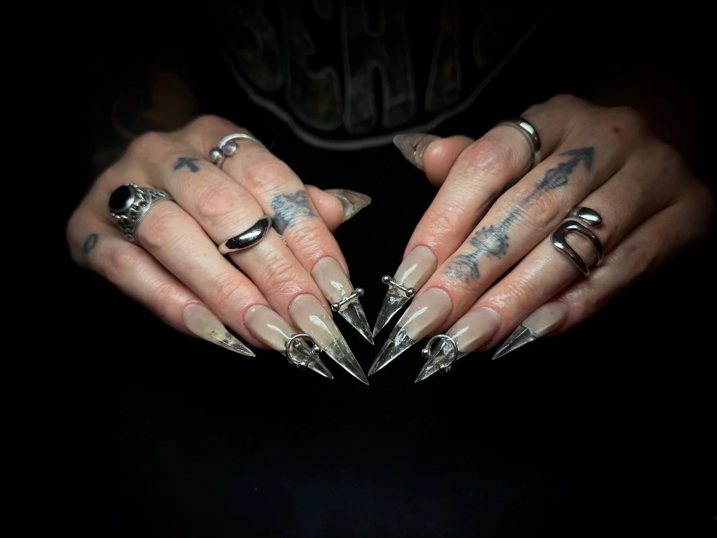 Close-up of hands with multiple tattoos and rings, displaying long, pointed acrylic nails with metallic accents against a black background.