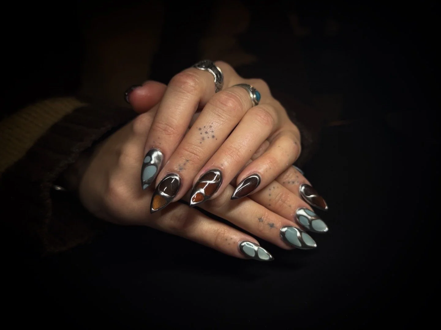 Close-up of hands with manicured nails featuring abstract, marbled designs in shades of brown, black, gray, and white, adorned with several rings.
