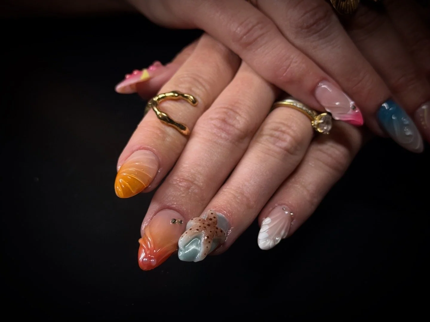 Close-up of hands with decorated nails in pastel colors and gold rings, dark background.