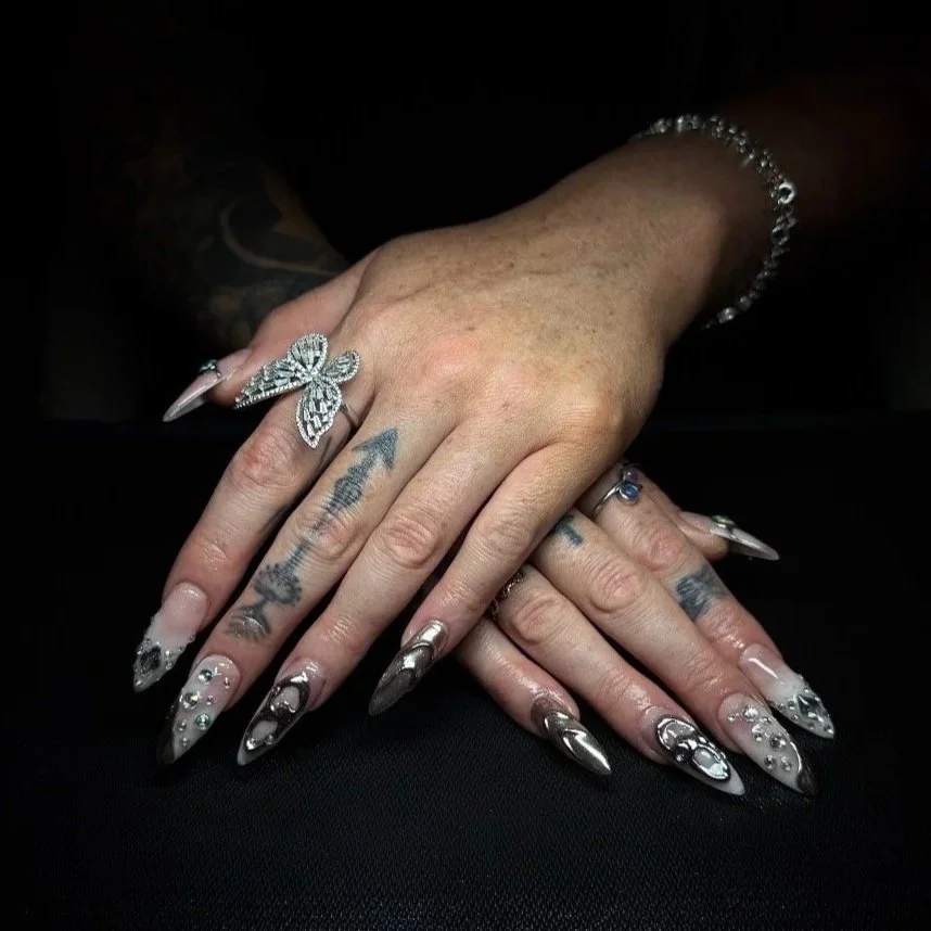 A person's hands with intricate silver and black nail art, tattoos, and jewelry, resting on a black surface.