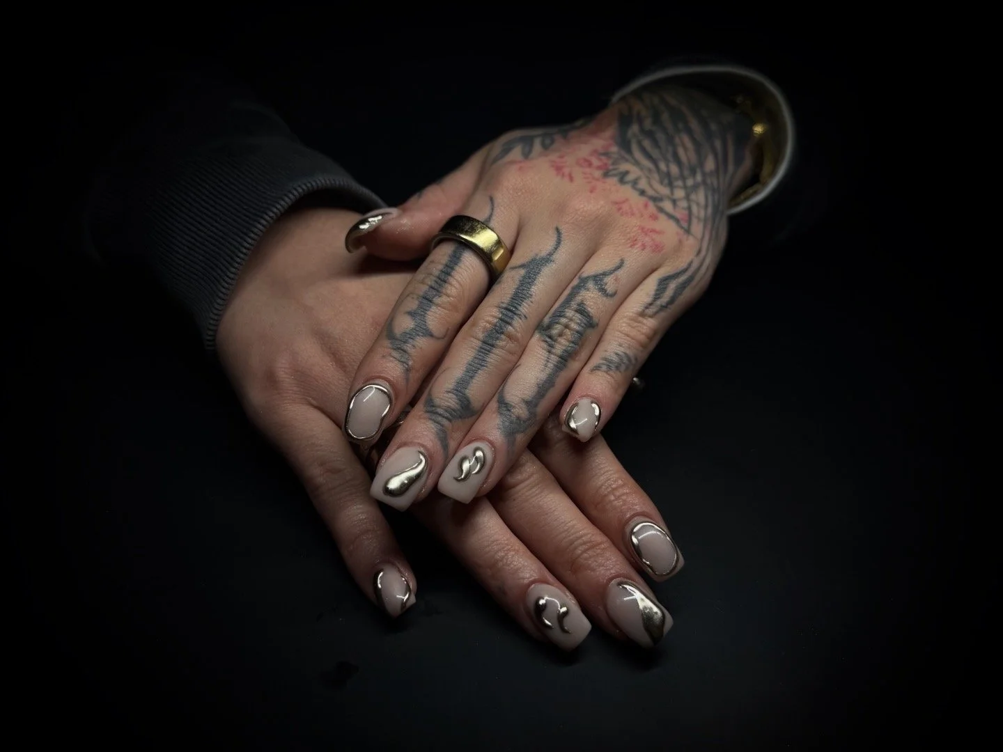 Close-up of two hands with tattoos, rings, and silver nail polish, resting on a black surface.