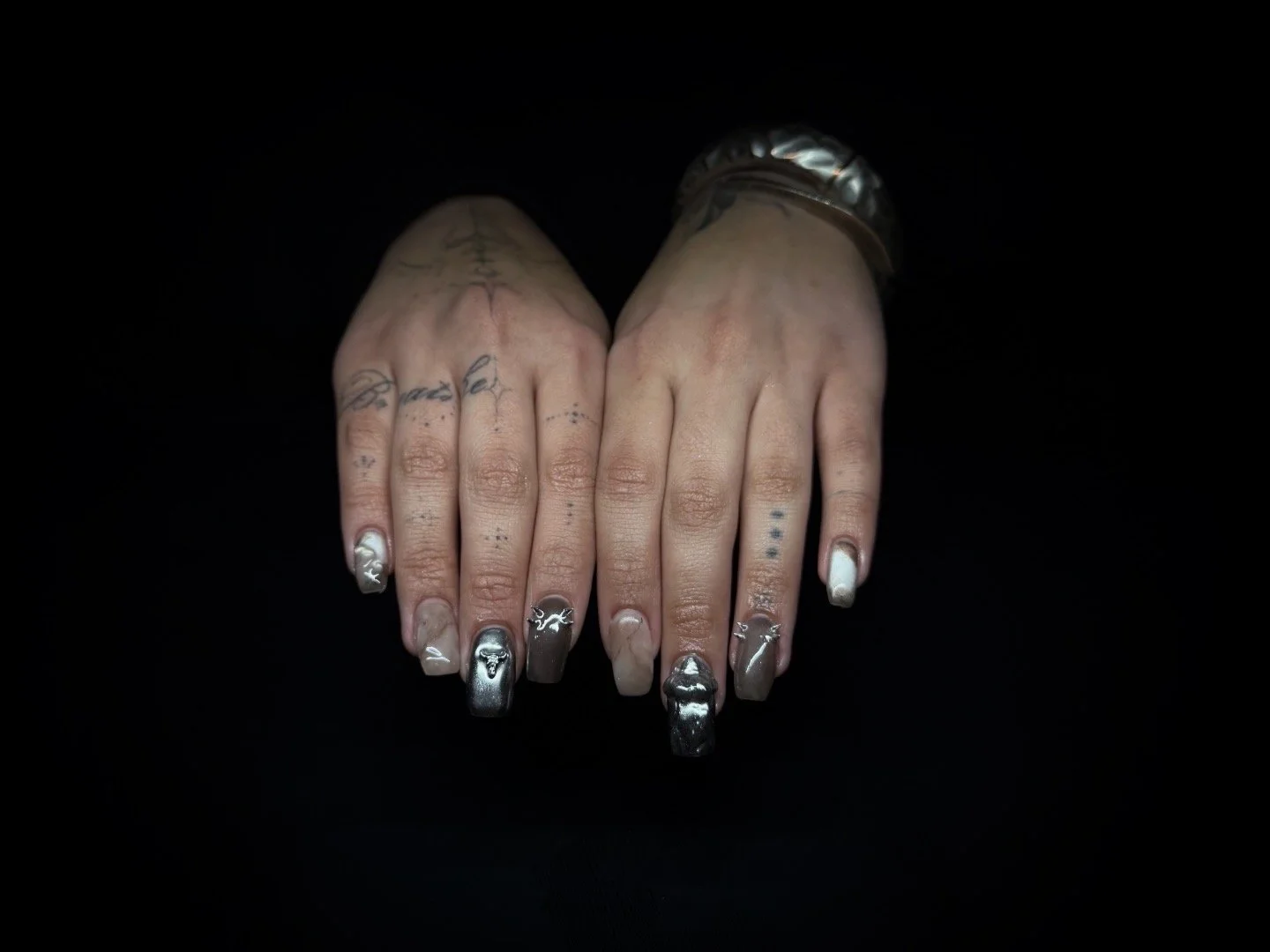 Two hands with tattoos and metallic silver and black nail polish against a black background.