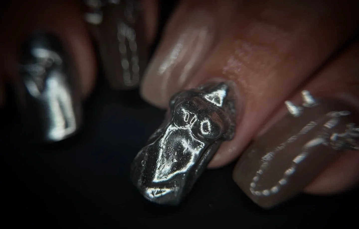Close-up of a finger with metallic nail art, featuring a 3D silver design resembling a futuristic or sci-fi theme.