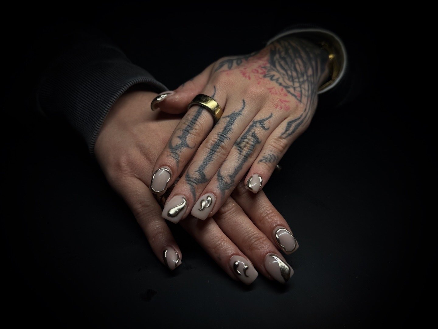 Two hands with tattoos and manicured nails resting on a black surface, one hand overlapping the other, adorned with rings and a bracelet.