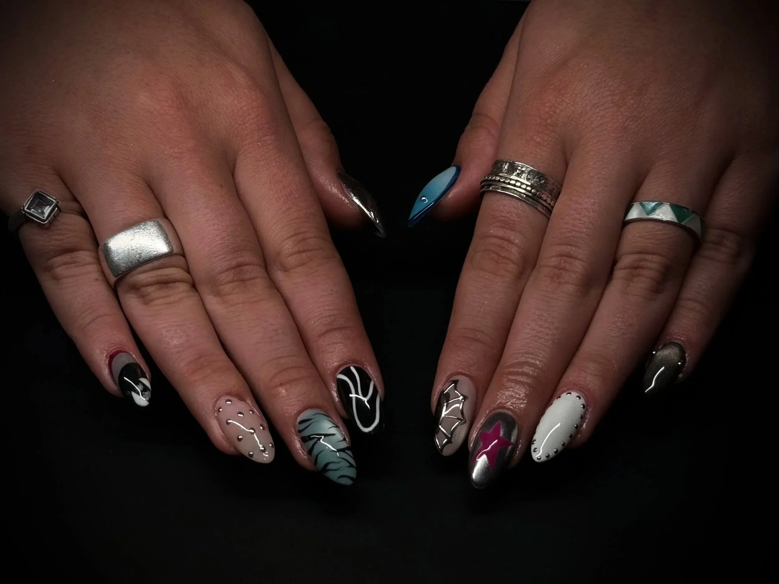 Close-up of hands with decorated nails and multiple rings on fingers, set against a black background.